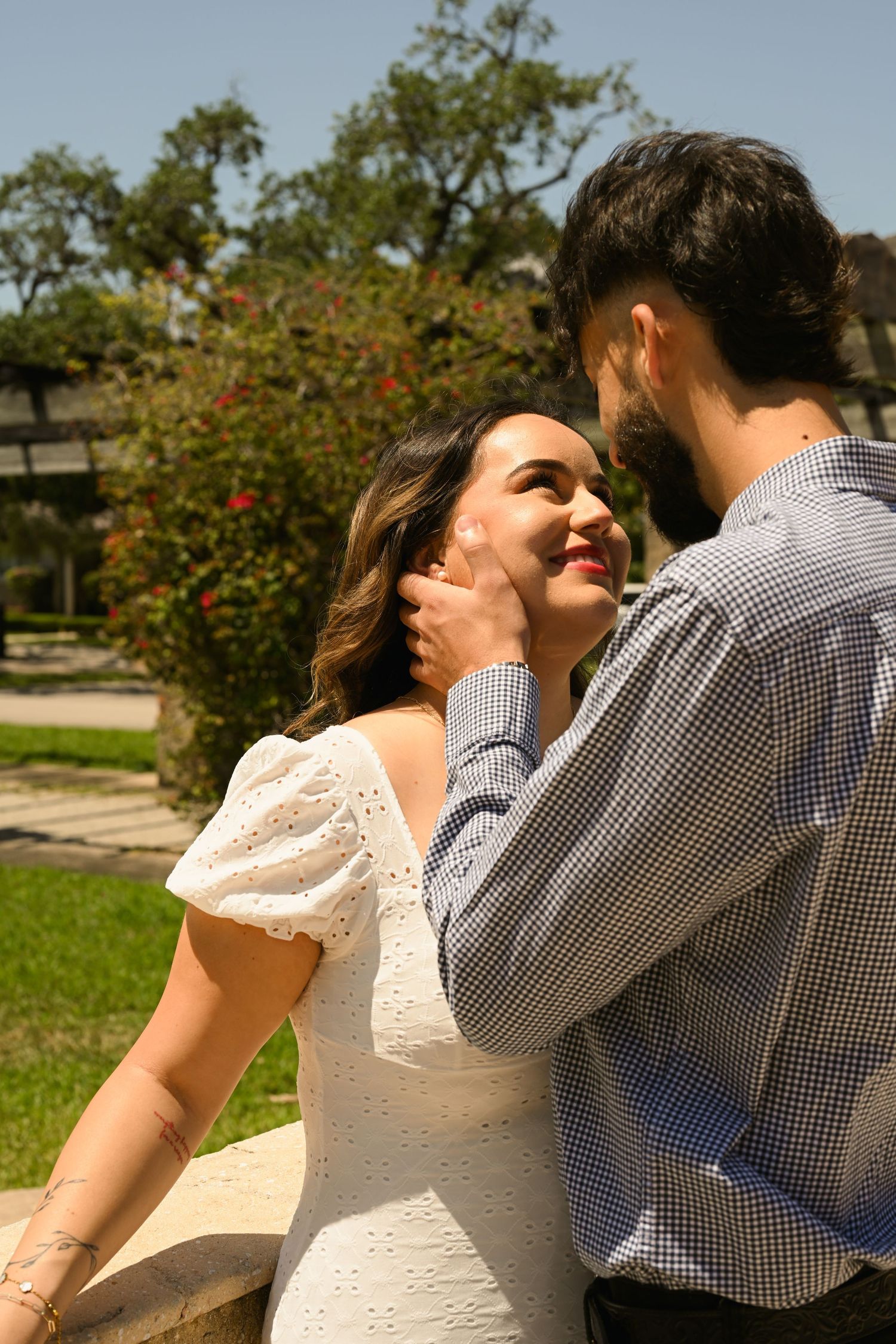 Romantic couple in white dress and checkered shirt share tender moments in a sunny garden setting.