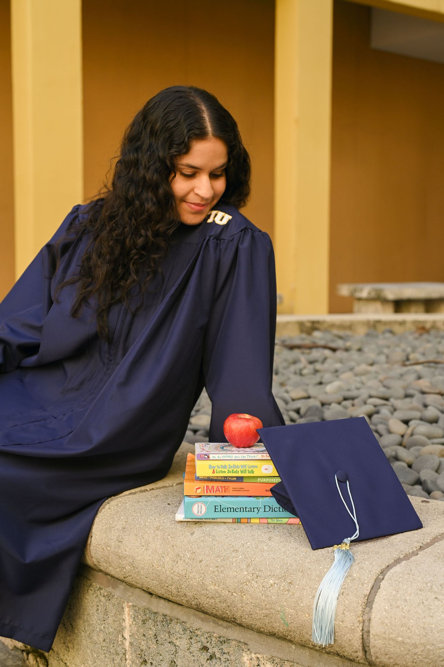 Graduate in navy gown sits beside stack of books and graduation cap.