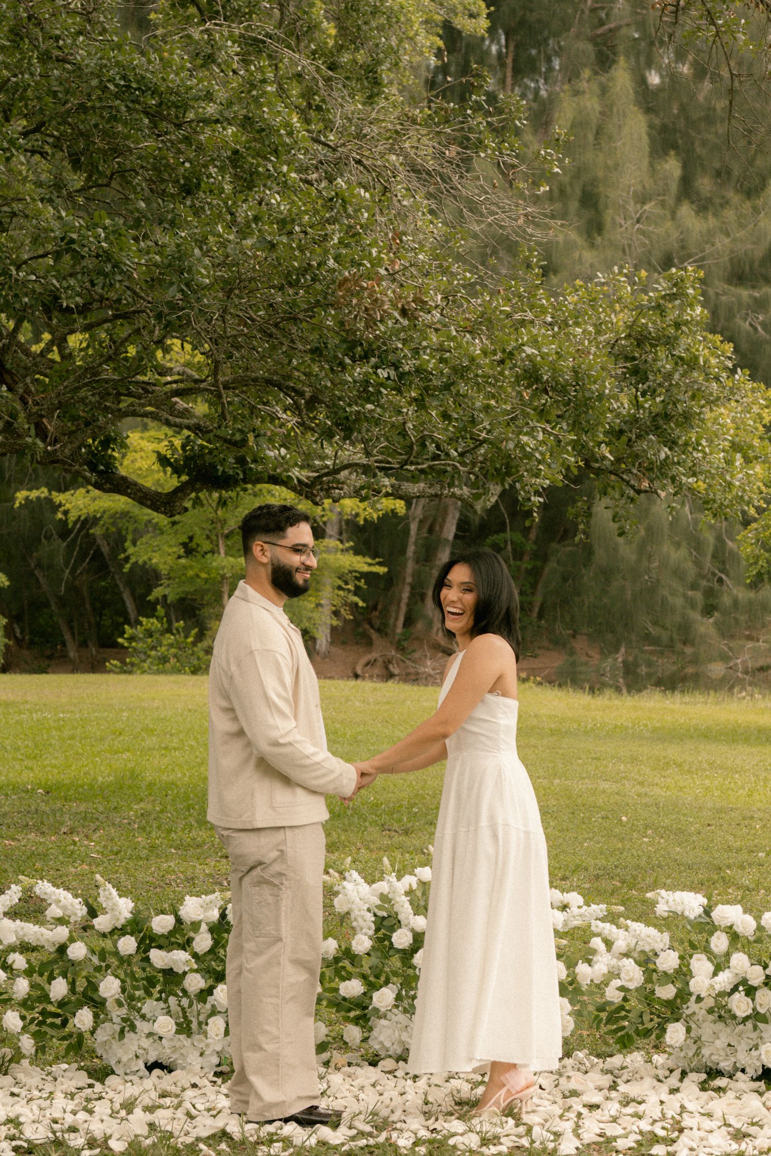 Couple holding hands and smiling in a garden with white flowers and lush green trees in the background.
