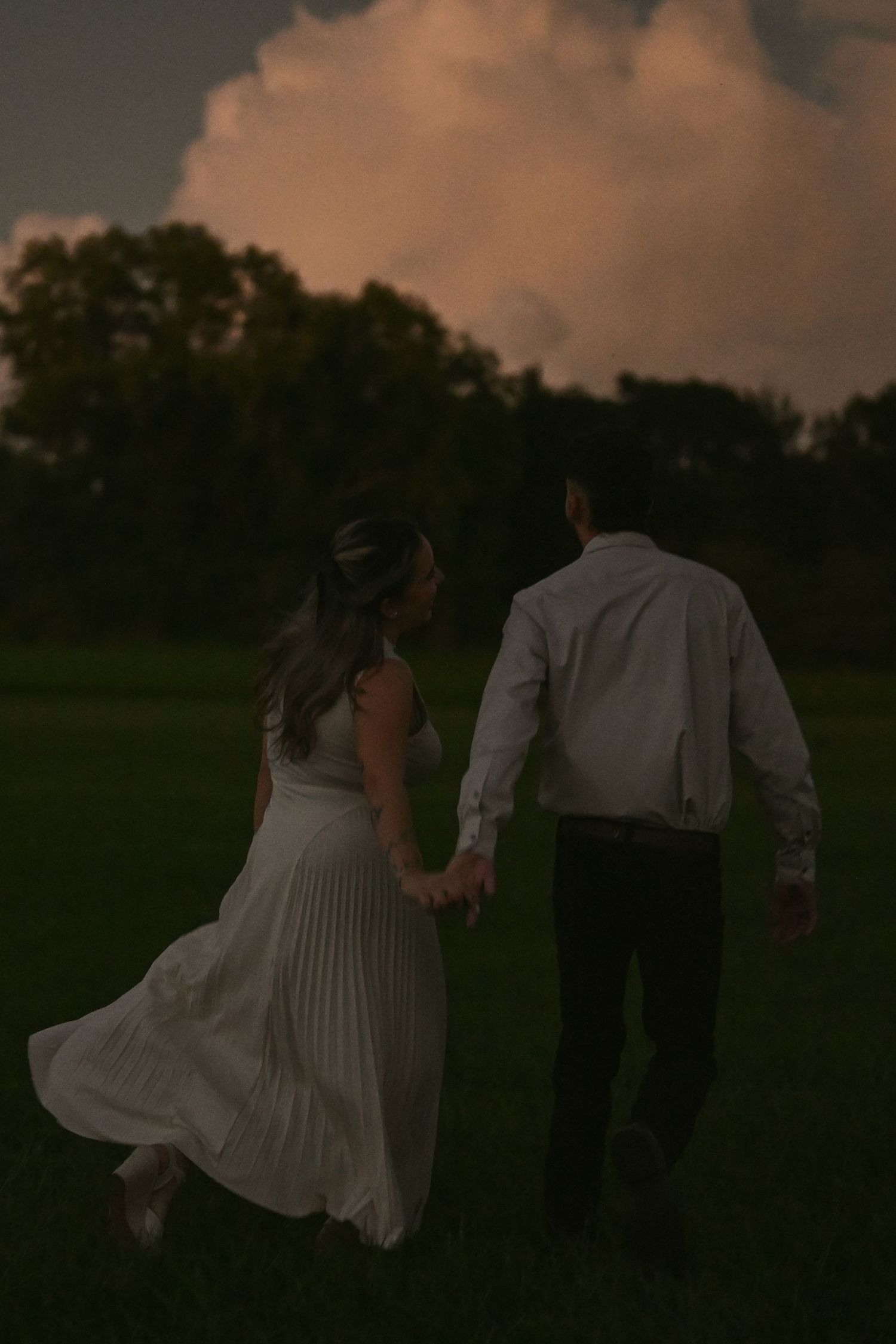 A couple in formal attire walks through a grassy field at sunset creating a romantic silhouette.