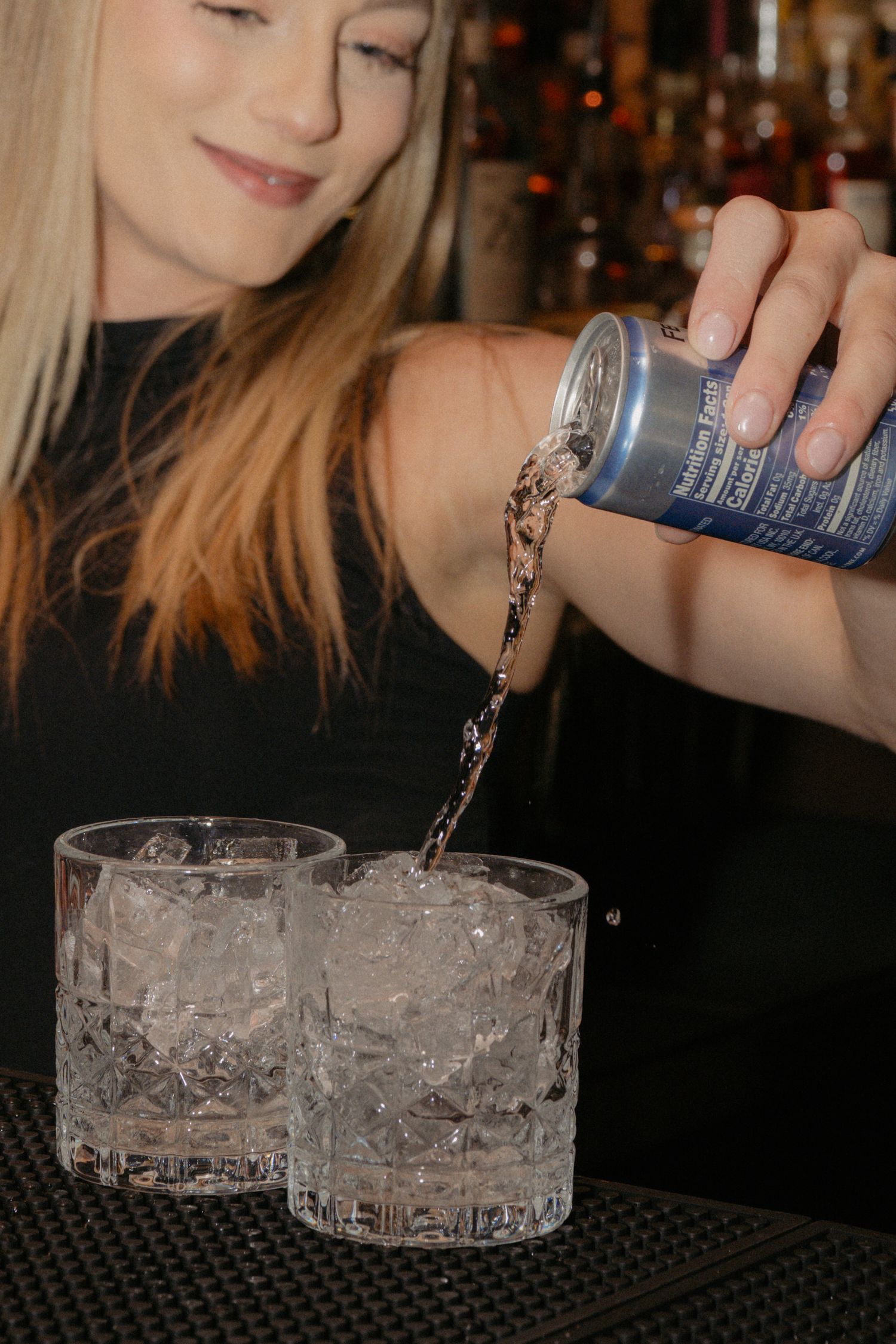 Blonde female bartender pouring a blue can drink over ice into crystal glasses at a bar.
