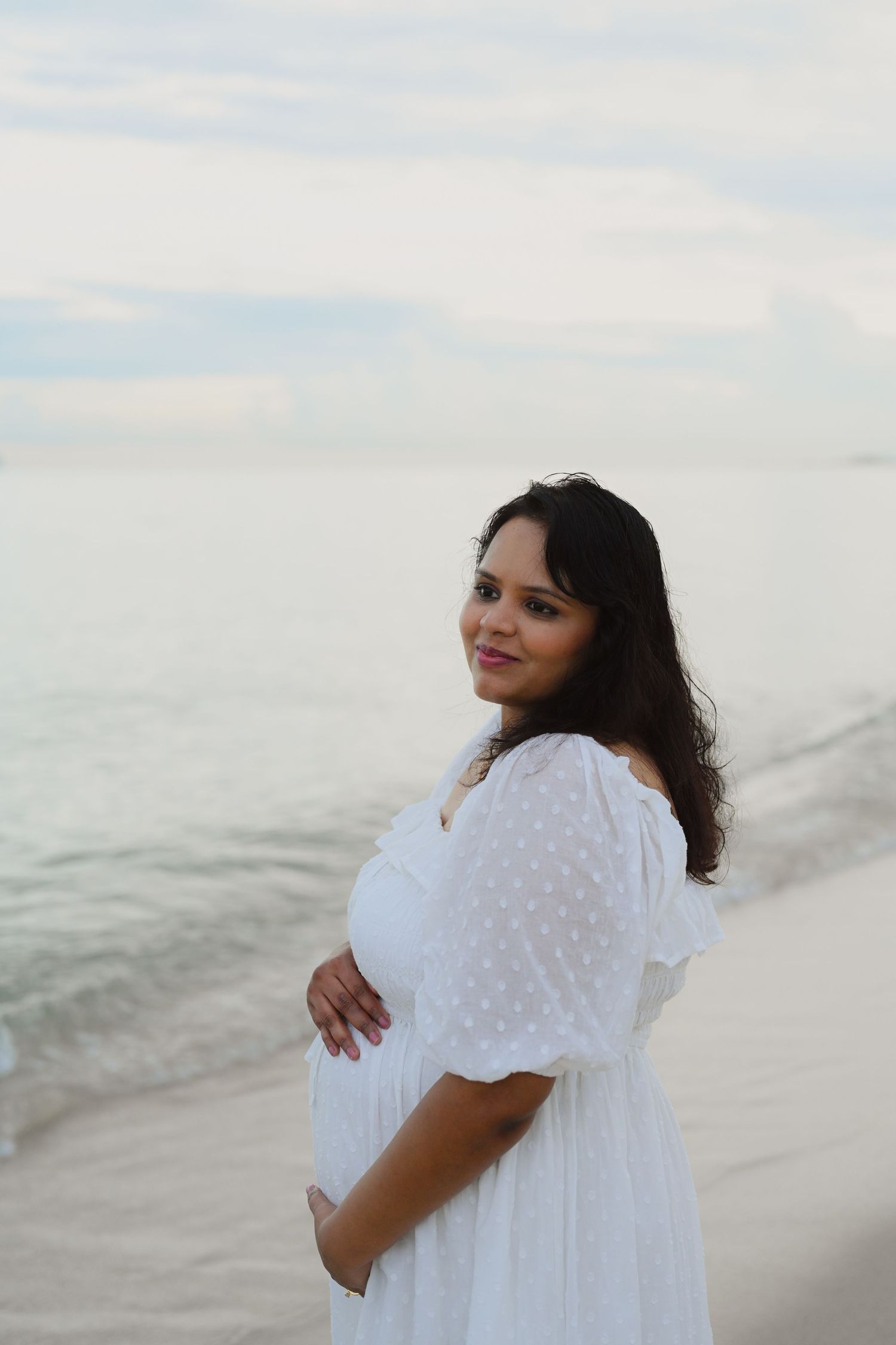 A person in a flowing white dress poses on a serene beach at sunset with waves in the background.