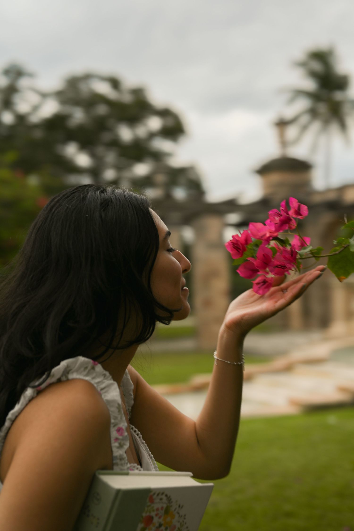 Someone in a white dress holds bright pink flowers against a backdrop of tropical architecture.
