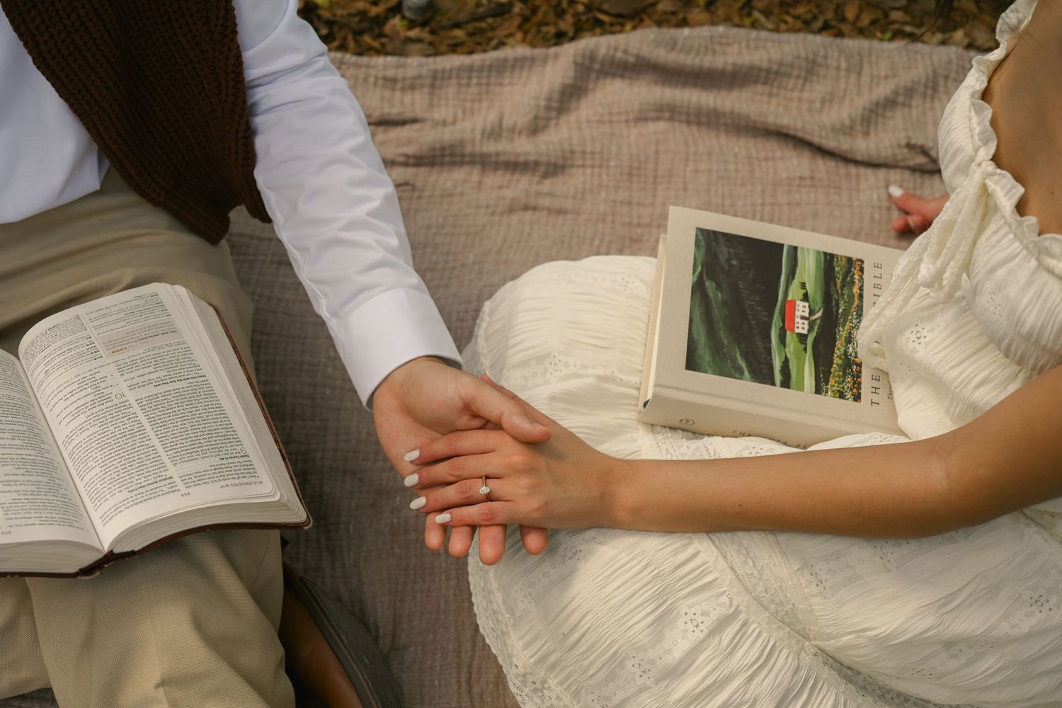 Newlywed couple holding hands on a blanket with an open Bible and book during an outdoor wedding picnic.