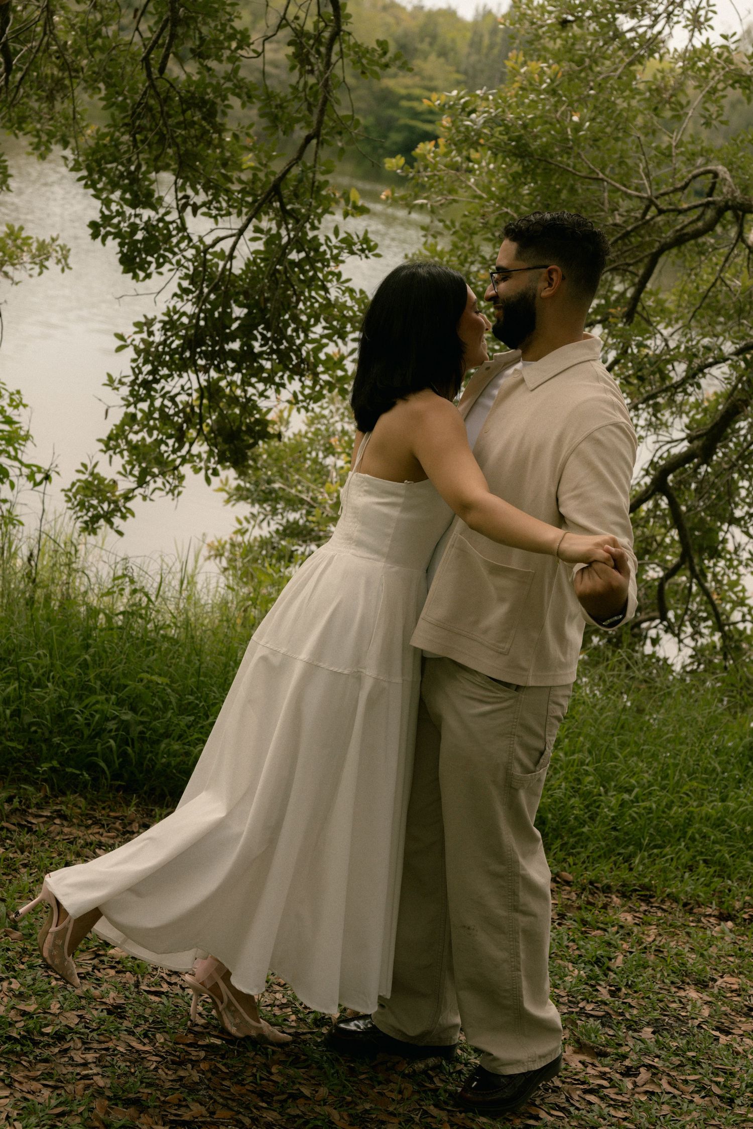 Couple embracing near a serene river, woman in white dress and man in beige outfit, surrounded by lush green trees.
