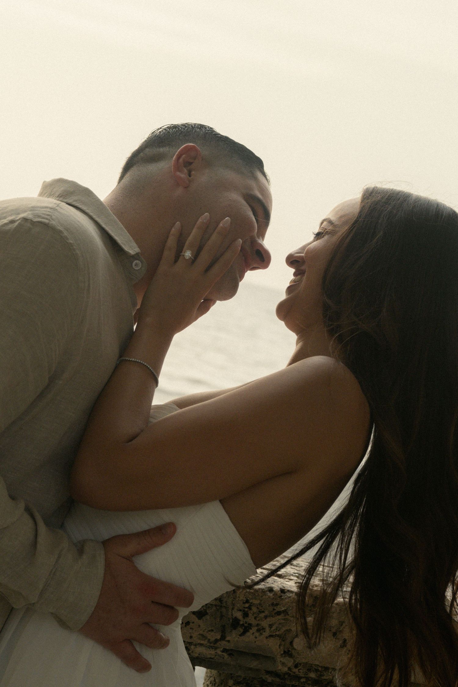 A couple shares a romantic moment, embracing and smiling at each other on a beach at sunset.