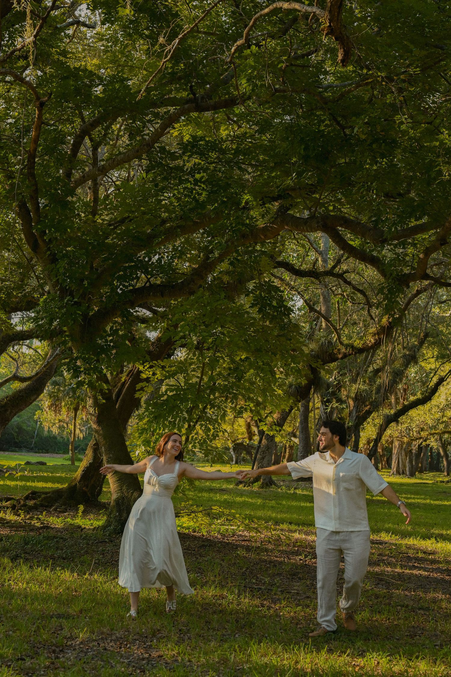 A couple dances joyfully under majestic oak trees draped in Spanish moss during golden hour in a serene park setting.