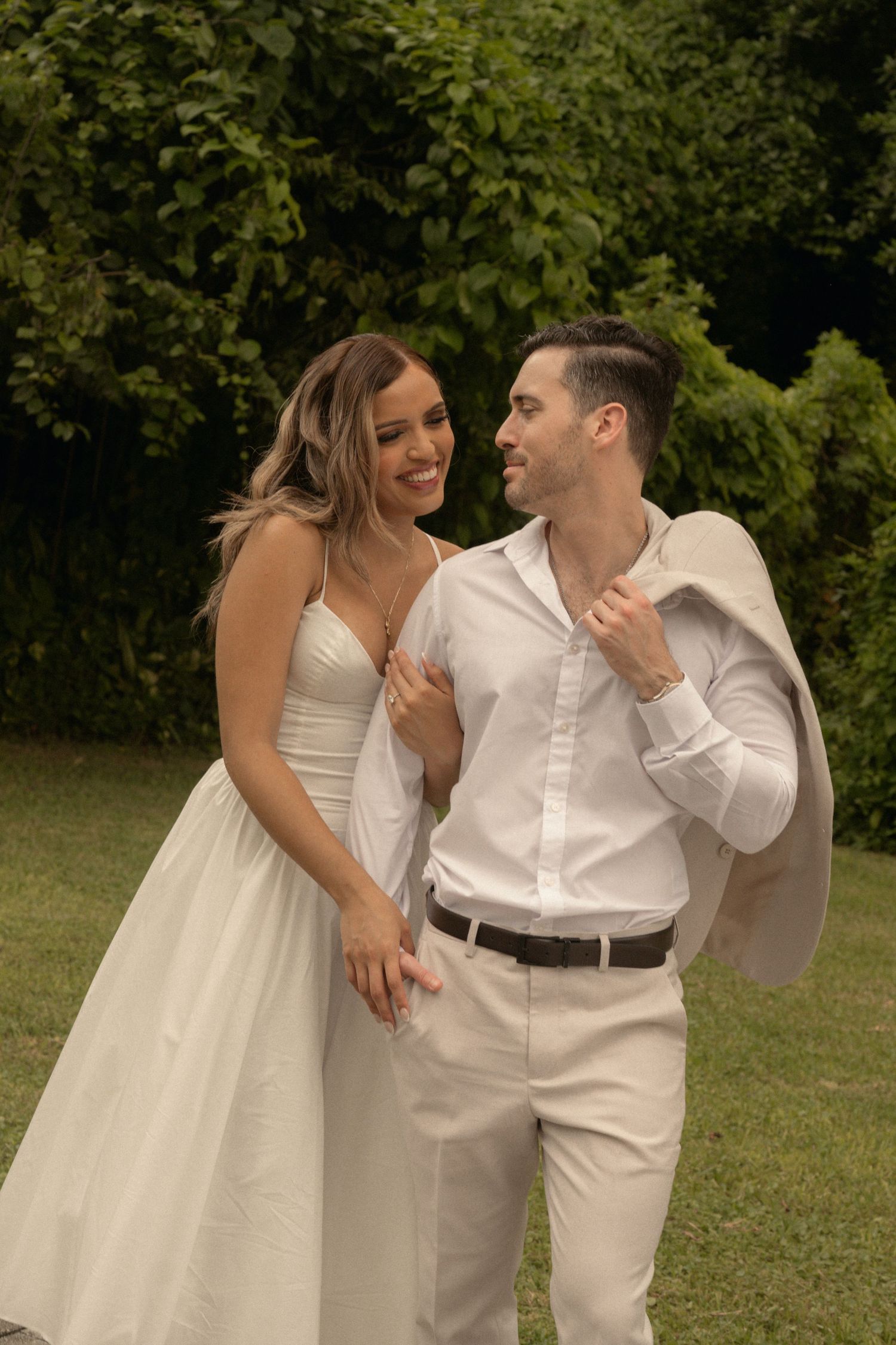 Smiling bride in a white dress embraces her groom in a white shirt and beige trousers on a lush green lawn at an outdoor engagement session.