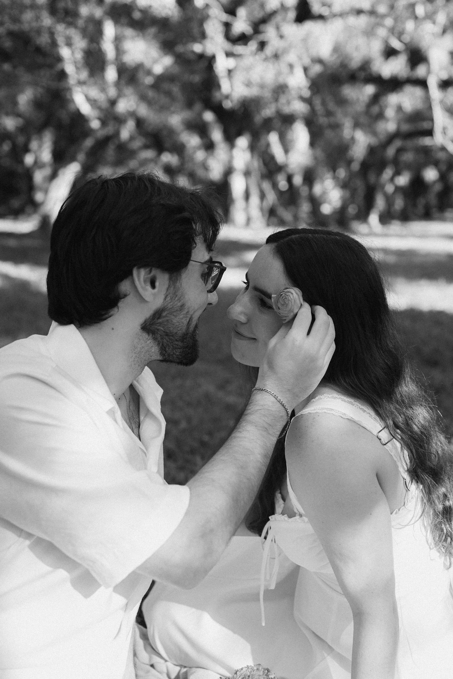 Black and white photo of a romantic couple almost kissing in a park, man gently touching womans hair.