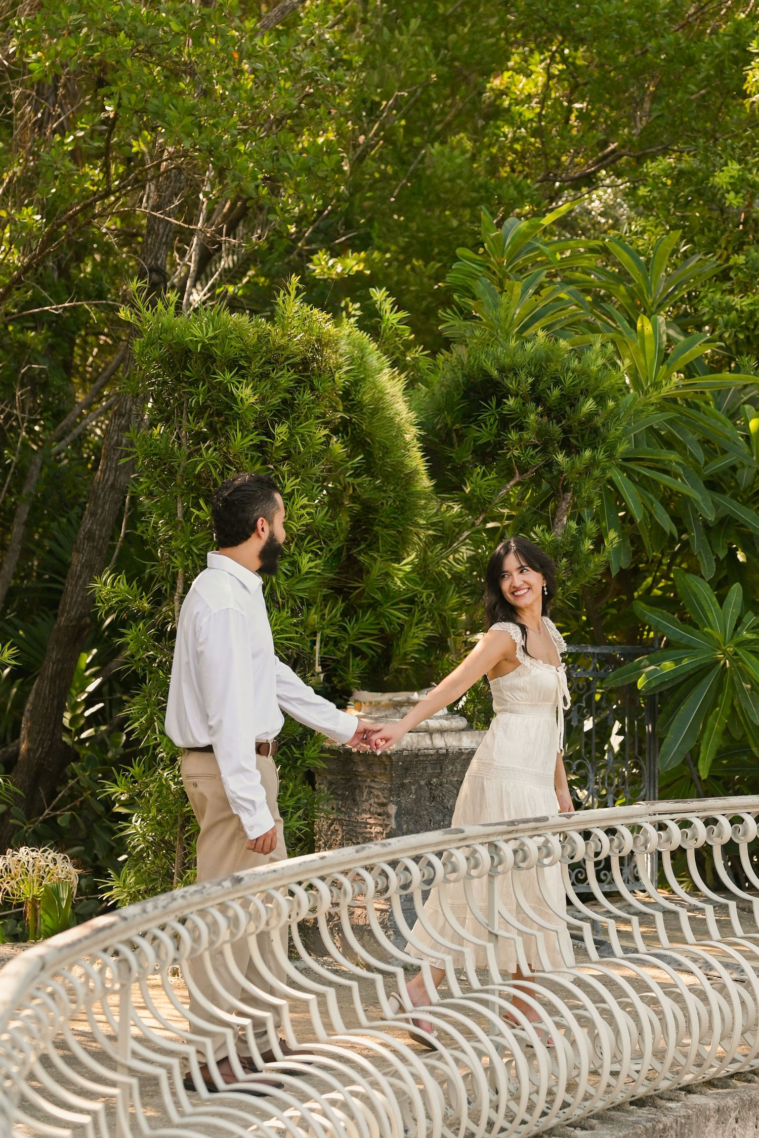 A couple walks along an ornate stone balustrade surrounded by tropical gardens and foliage.