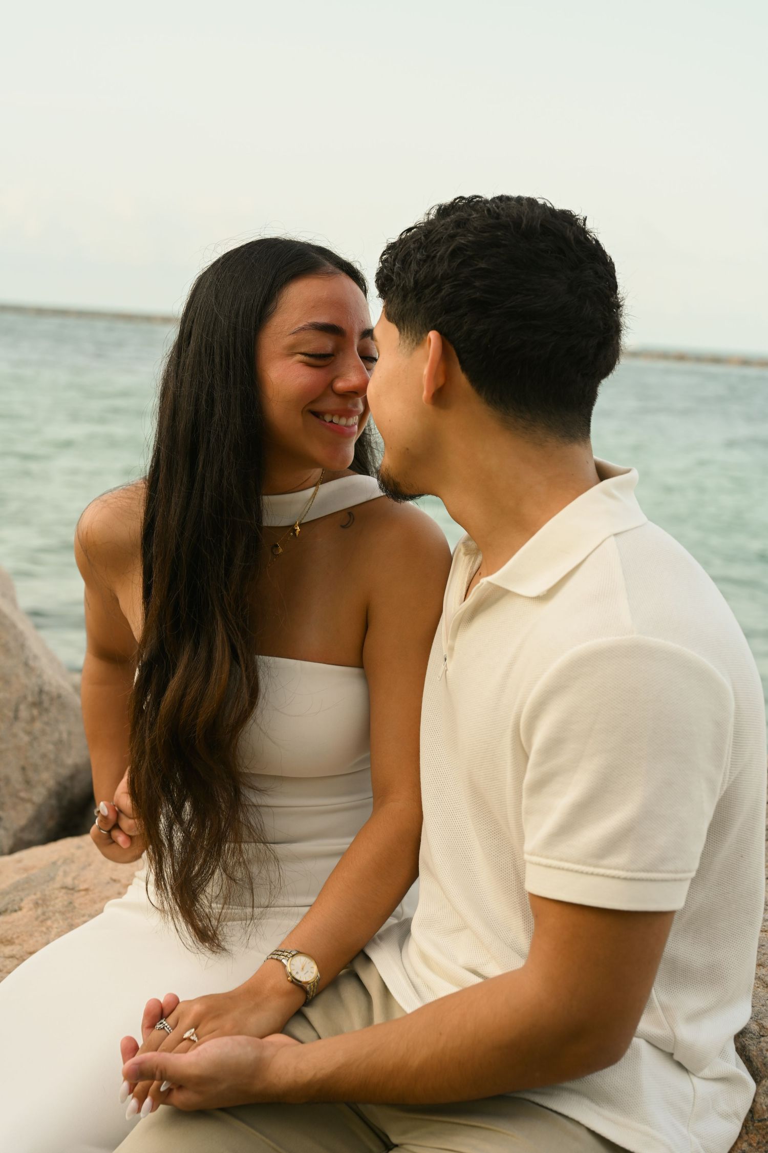 Intimate couple in white clothing sharing tender moments on rocky waterfront.