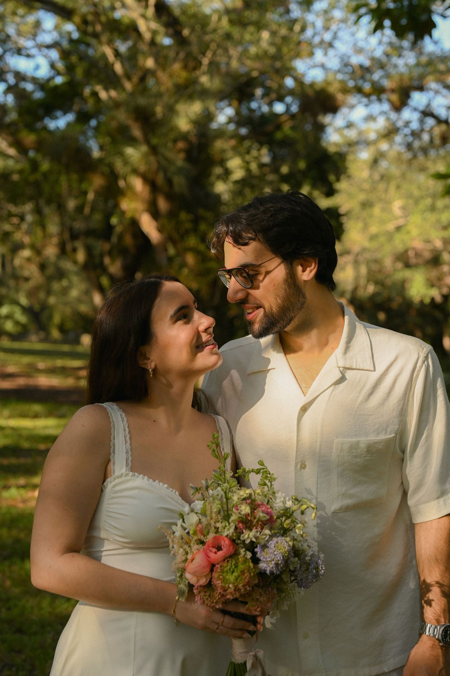 A romantic couple shares intimate moments in a sunlit park setting while holding a bouquet of fresh flowers.
