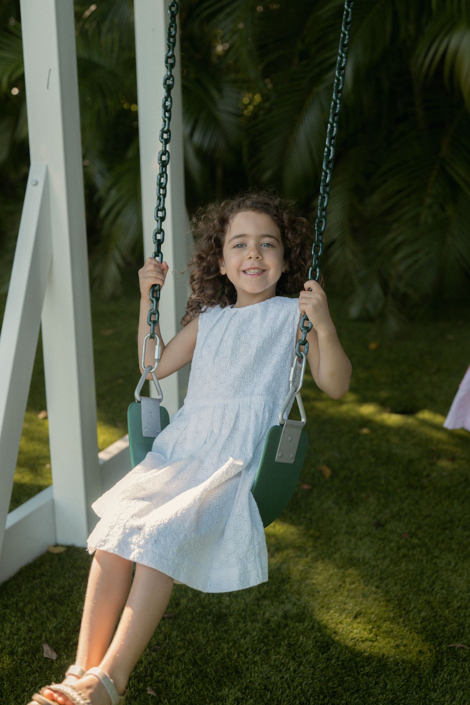 Smiling girl with curly hair in a white dress swings on a green swing set in a sunny garden surrounded by tropical plants.