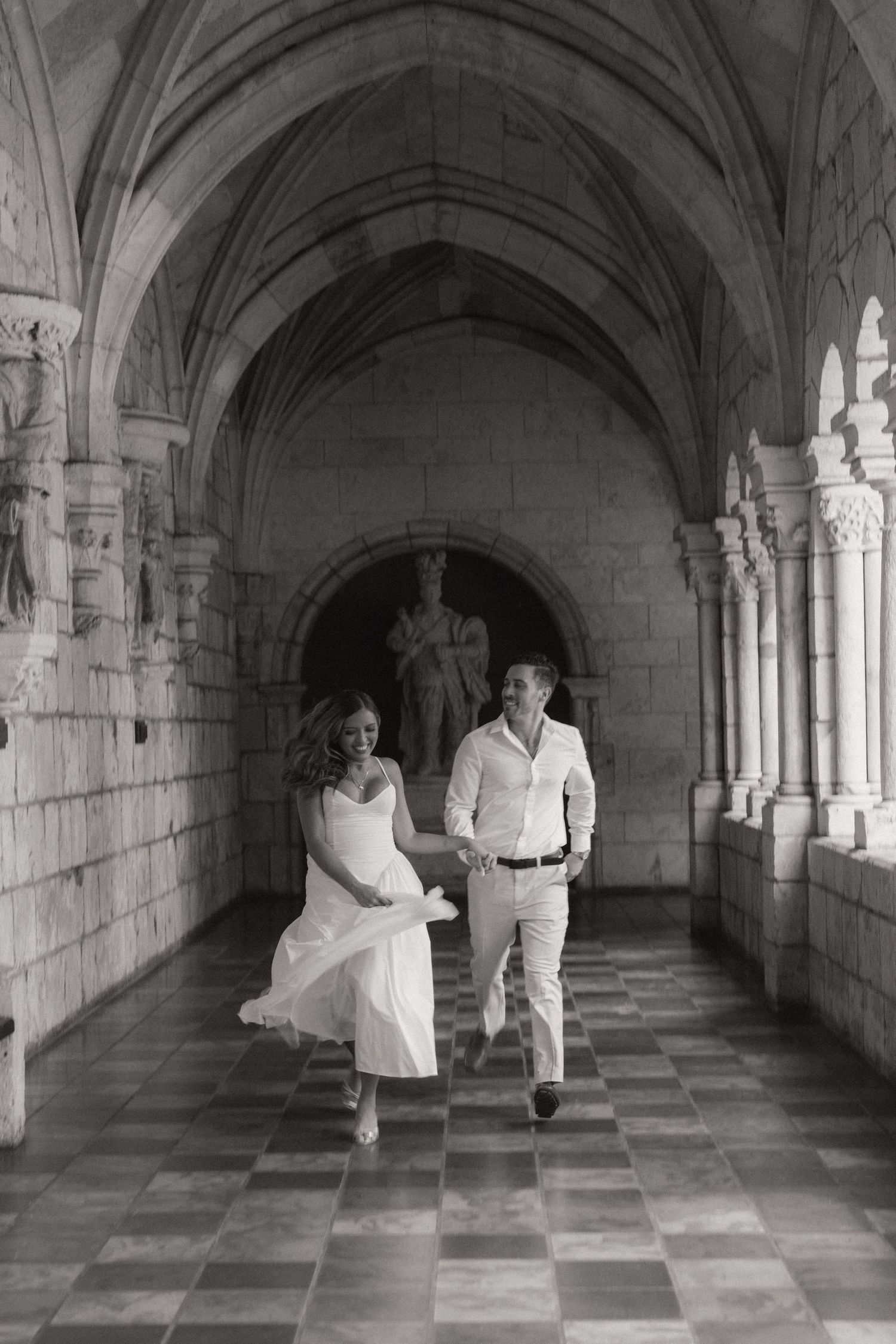 Black and white photo of a couple running hand in hand through a gothic stone cloister with a checkered tile floor.