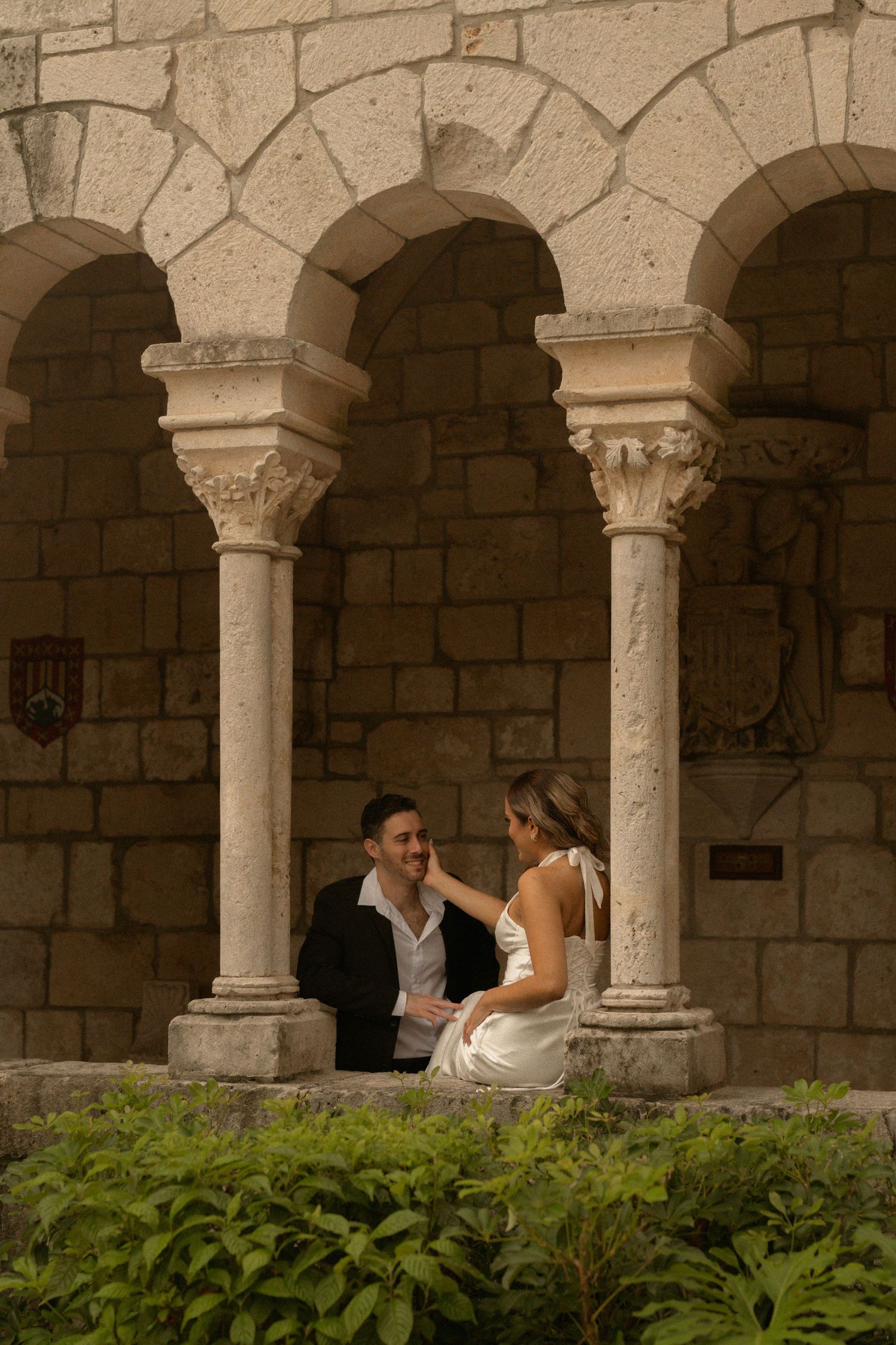 Engaged couple in elegant attire share a tender moment between stone arches and columns in a historic courtyard surrounded by greenery.