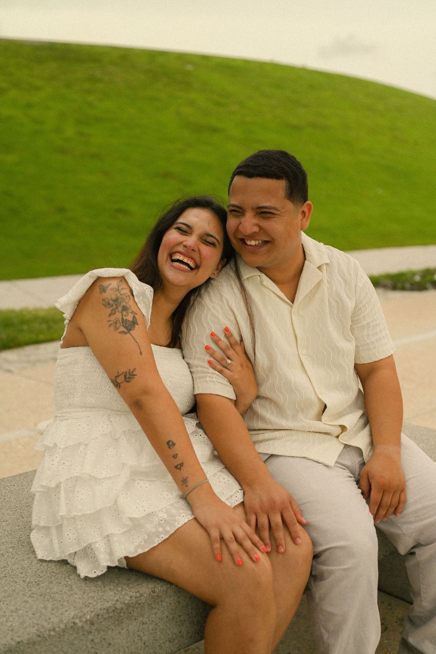 A couple in light colored outfits share a candid laughing moment while sitting outdoors.