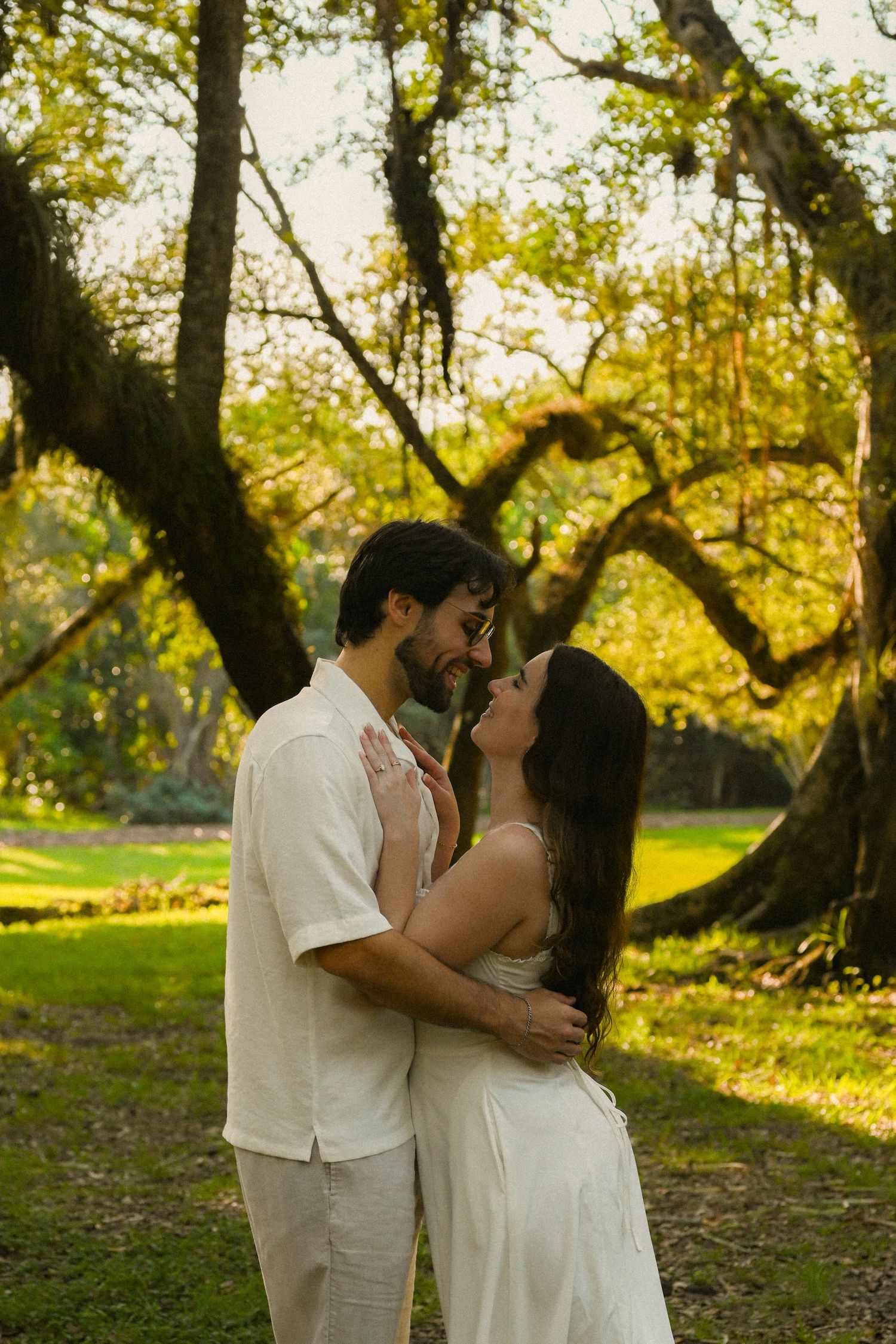 A couple in white attire shares a romantic moment under a large tree in a sunlit park.