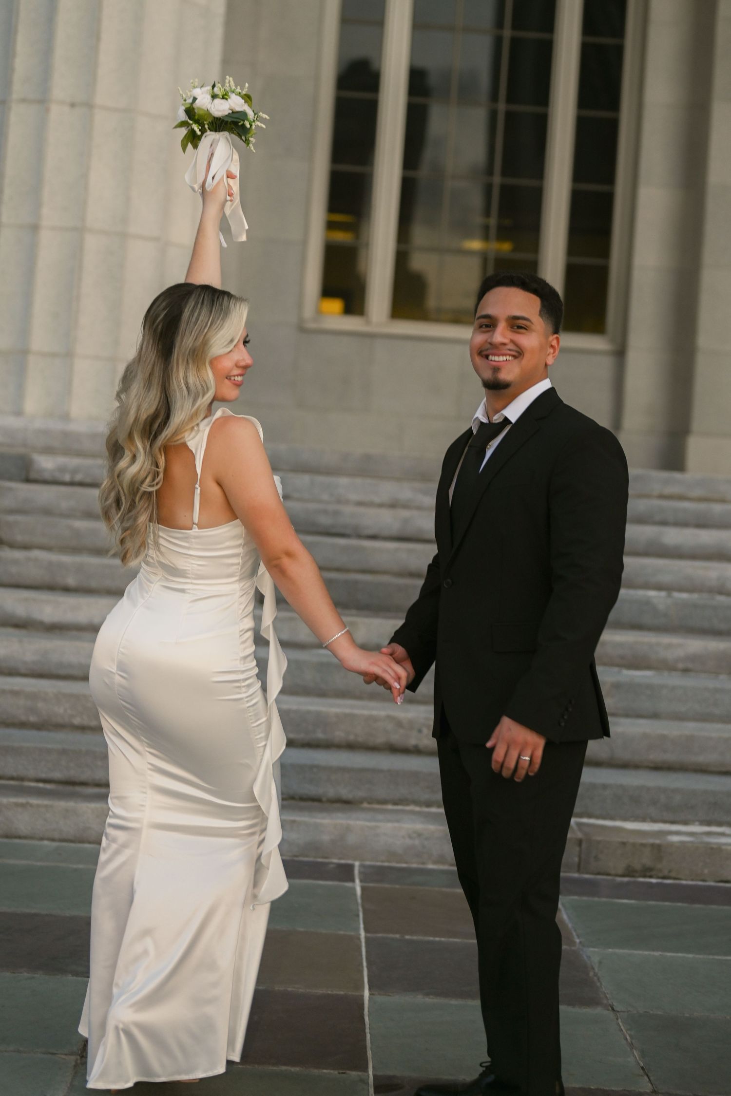 A bride in a white dress and groom in a black suit share a candid moment on steps with classical columns.