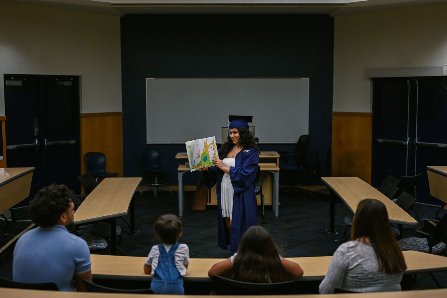 Graduate in navy regalia addresses audience in classroom lecture hall.