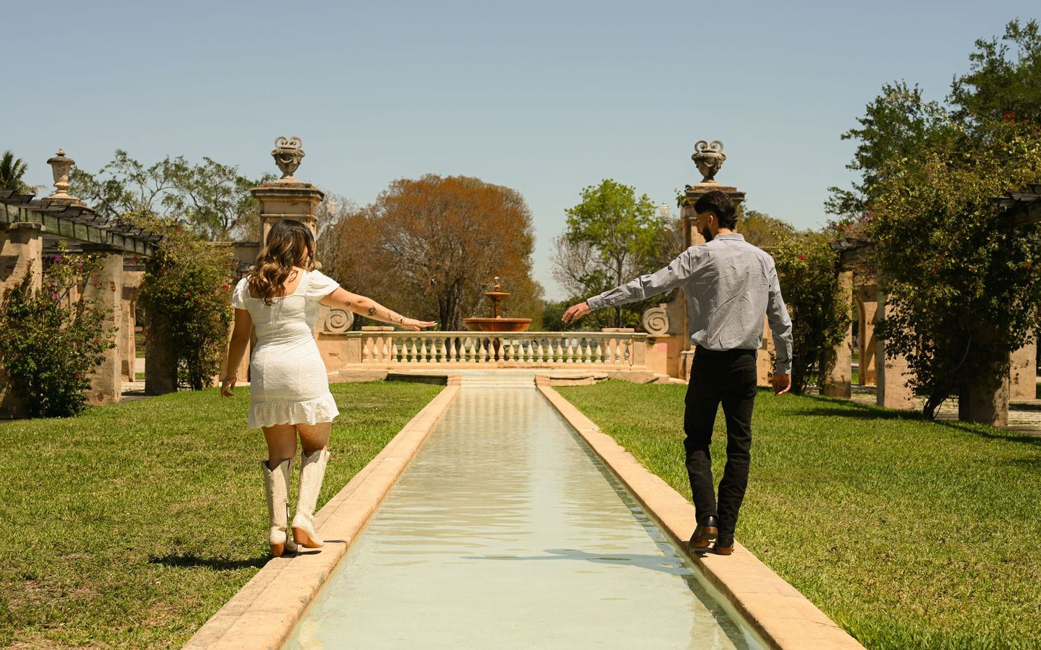 A romantic couple walks along a stone-lined path in a garden with ornate pillars and lush greenery.
