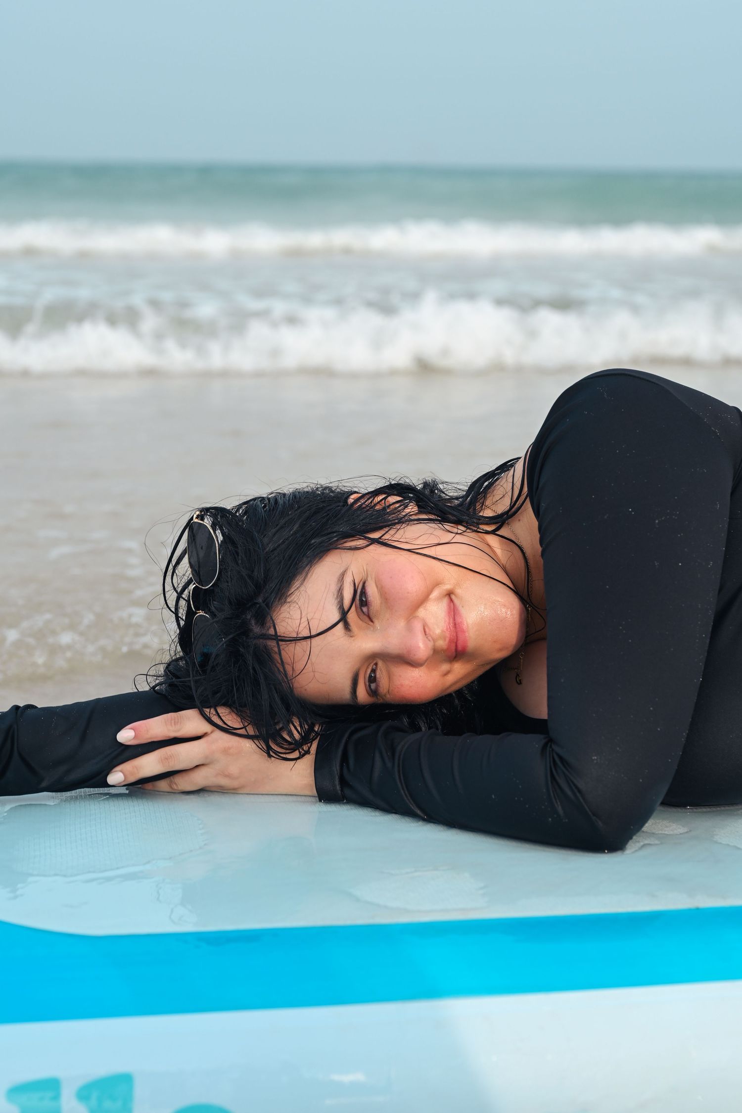 Person in black top lying on blue surfboard with wet hair at oceanside.