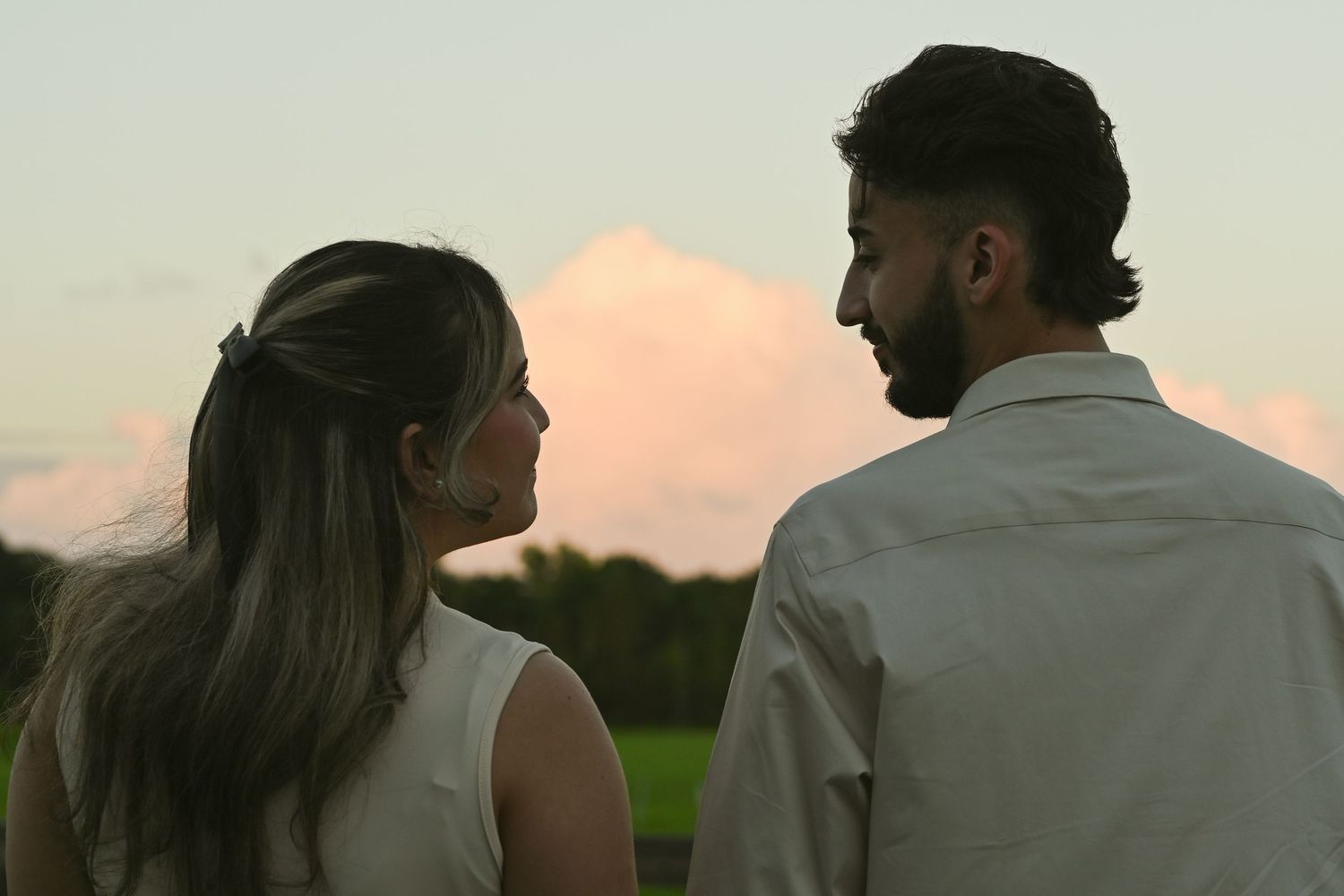 A couple gazes at each other against a pink sunset sky in a peaceful outdoor setting.