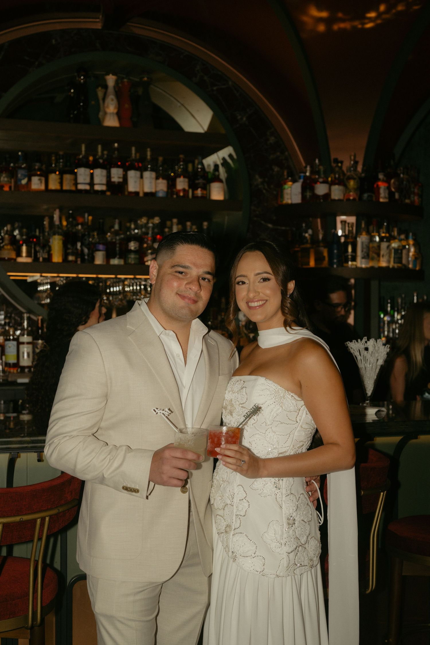 Bride and groom smiling together at a bar, holding cocktails, wearing white wedding attire.
