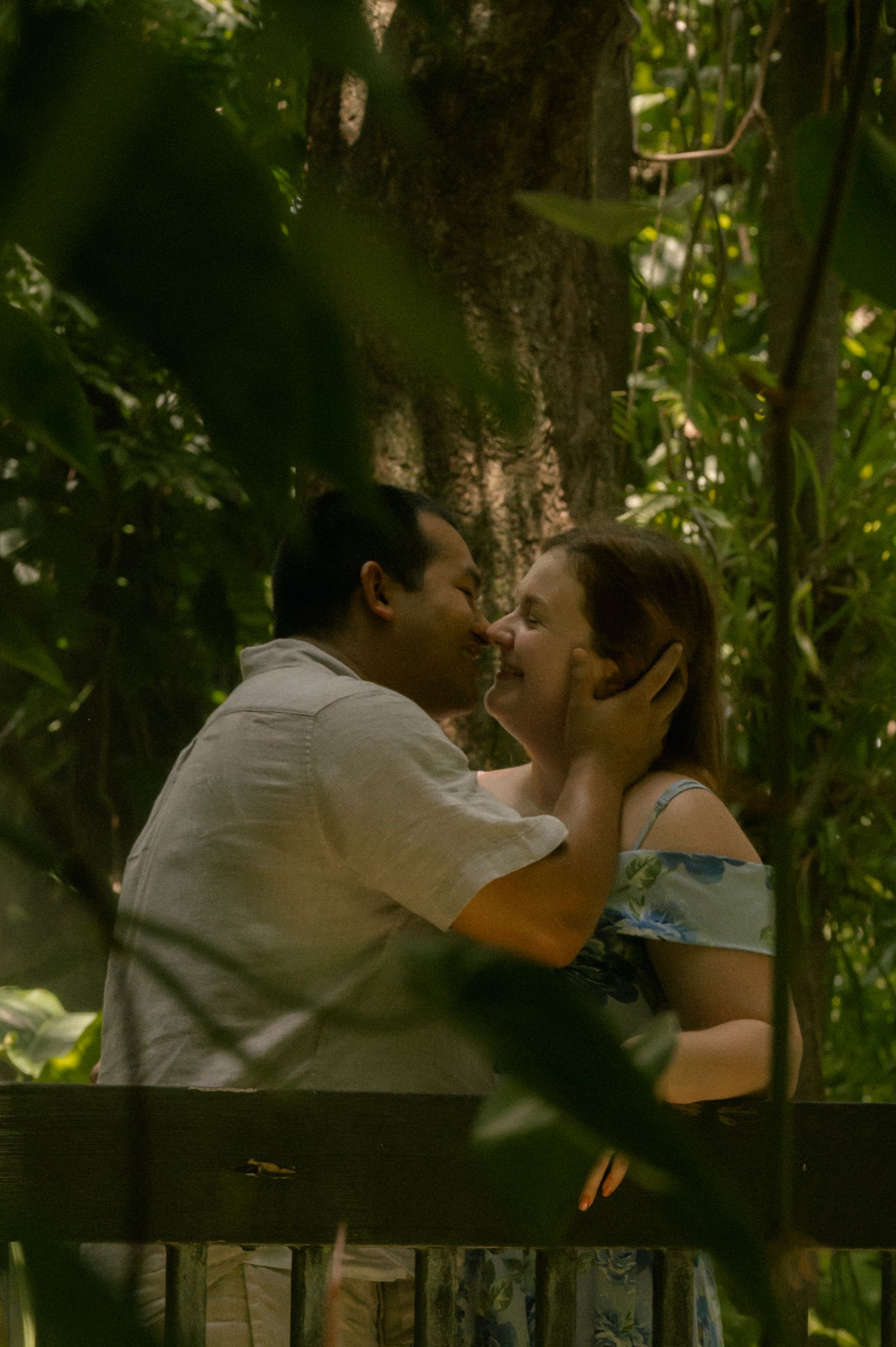 A couple shares a tender kiss on a bench surrounded by lush green foliage in a romantic outdoor setting.