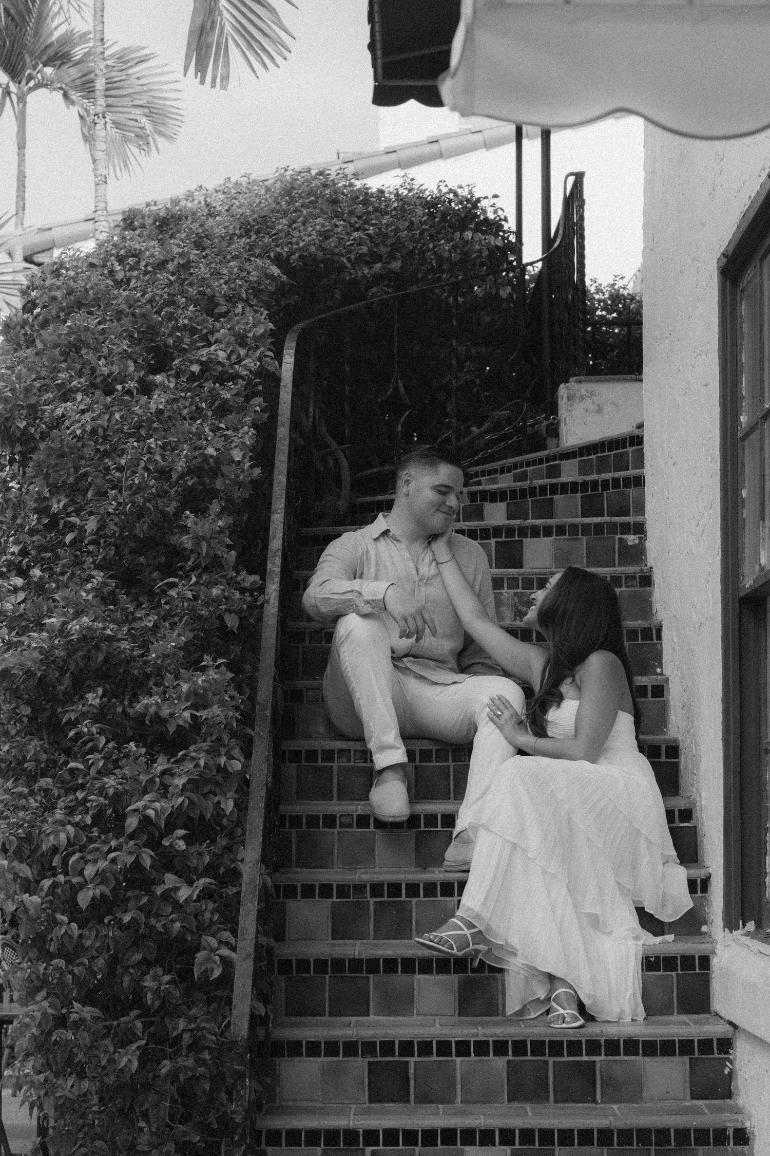Couple sitting on decorative tiled steps outside a building, surrounded by lush greenery, in black and white.