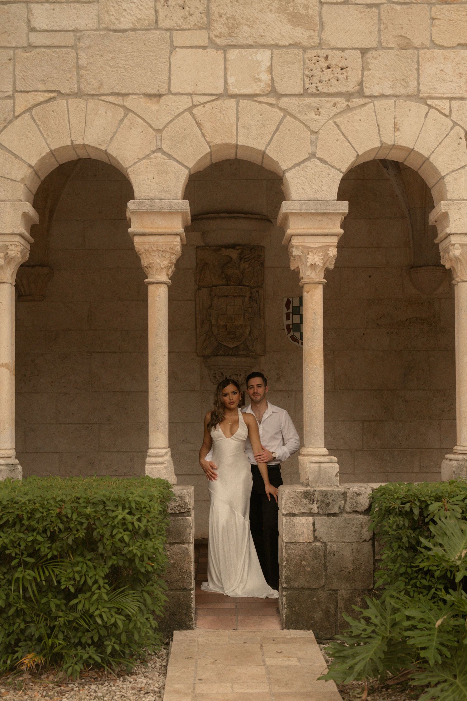 Engaged couple in elegant formal attire standing together beneath stone arches in a historic monastery garden.