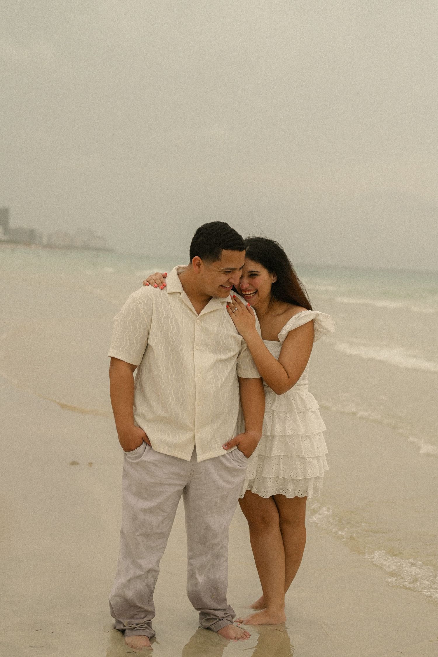 A couple in coordinated white outfits shares a quiet moment together on an overcast beach.