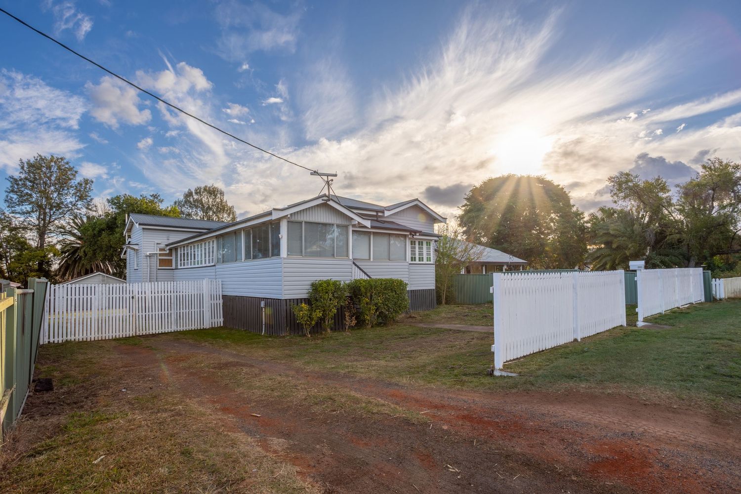 White Queensland weatherboard home with sunburst sky, white picket fence, and dirt driveway surrounded by lush green trees.