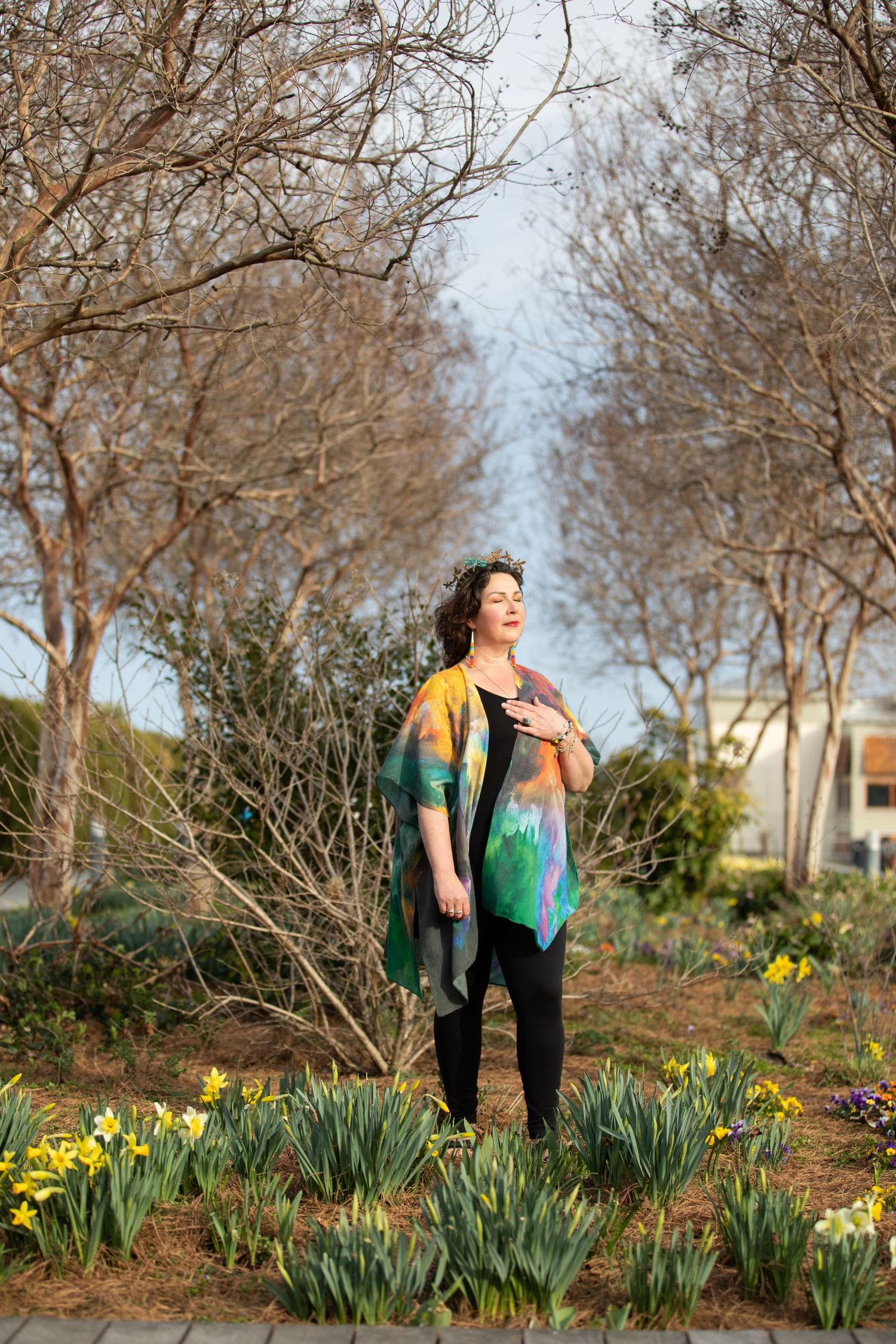 A person in a colorful yellow and teal dress stands among daffodils and bare trees in a spring garden setting.