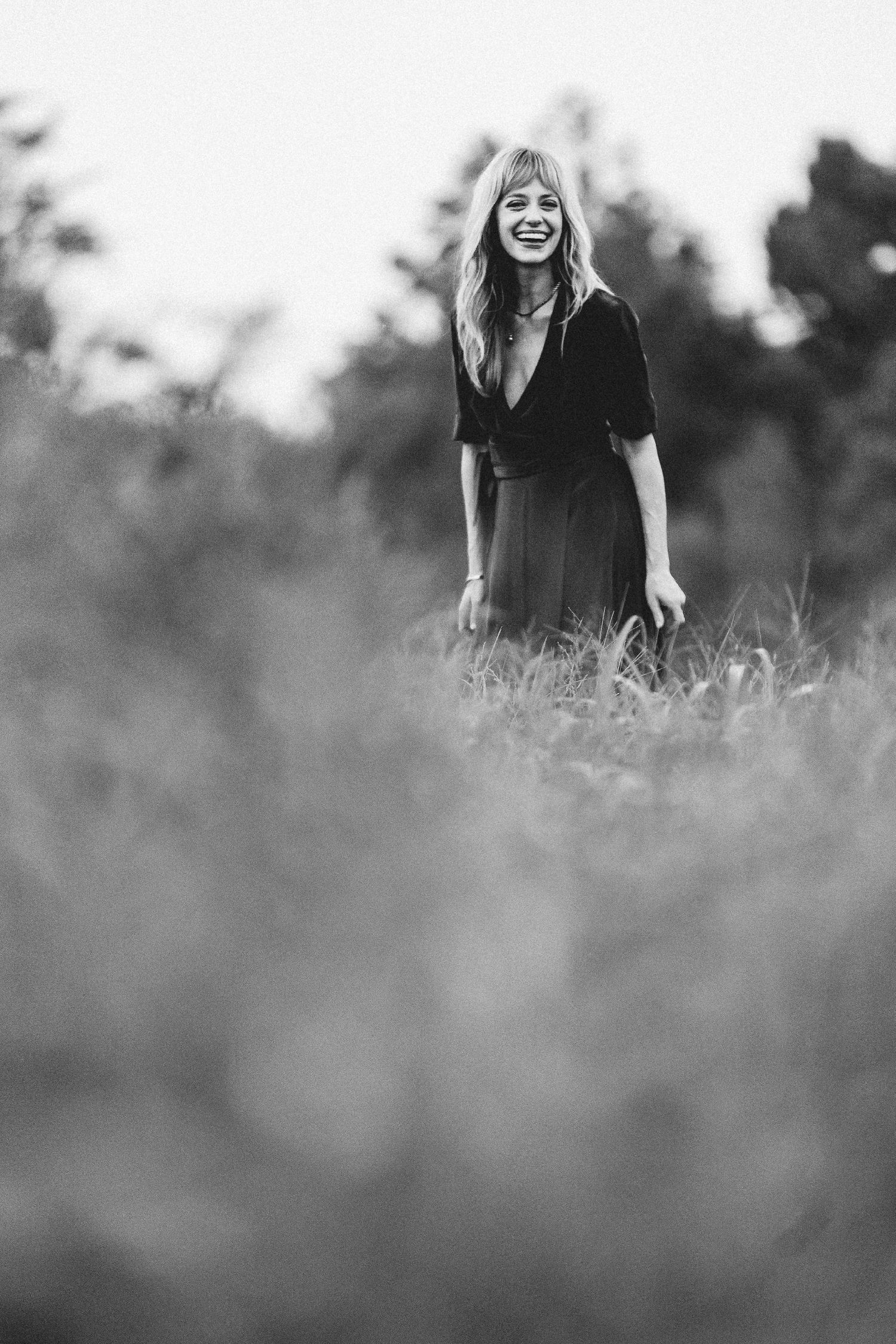 Black and white photo of a smiling woman in a dress standing in a field with trees in the background.