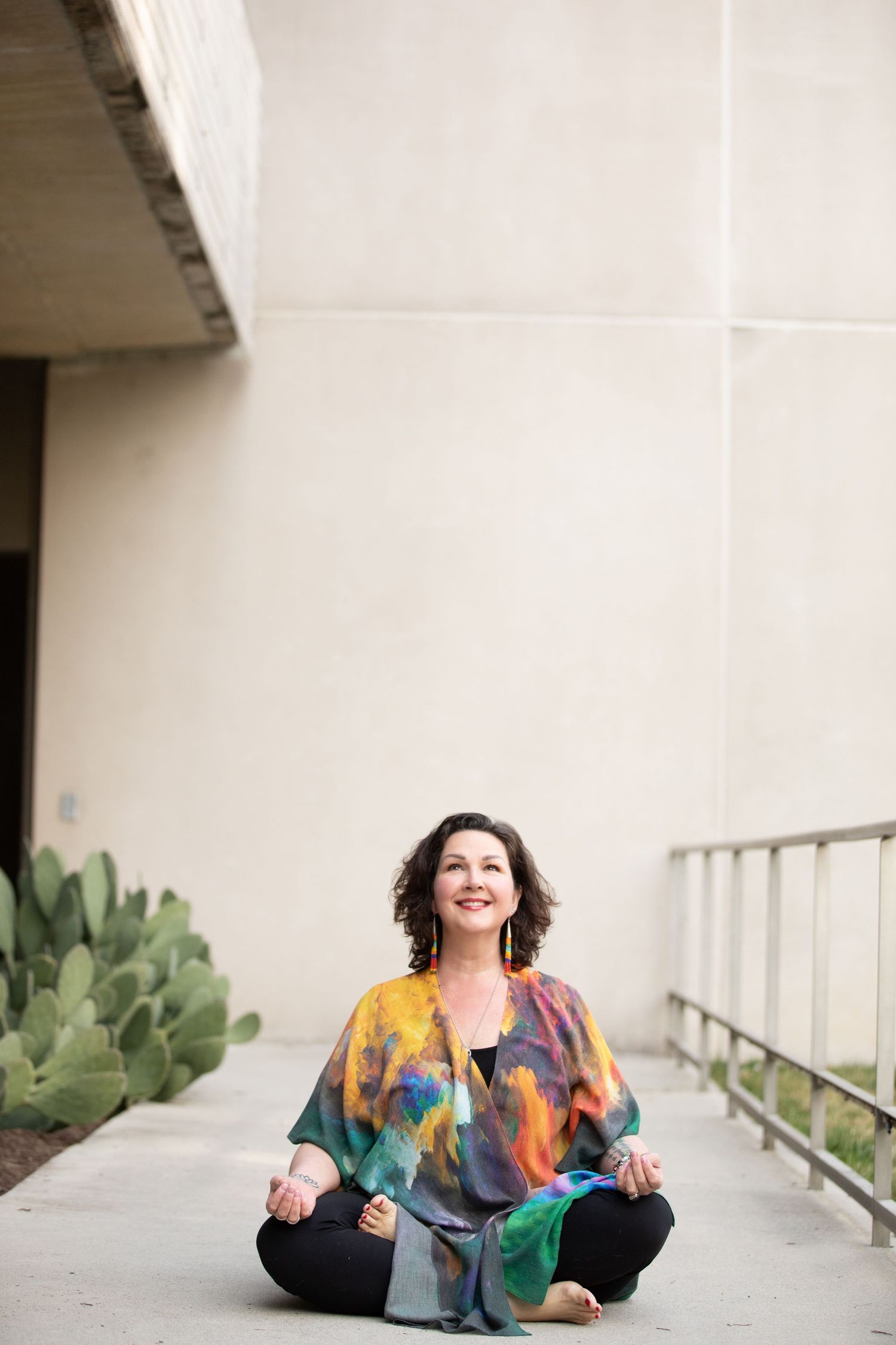 A colorfully dressed person sits in a lotus meditation pose on a concrete walkway with cactus plants nearby.