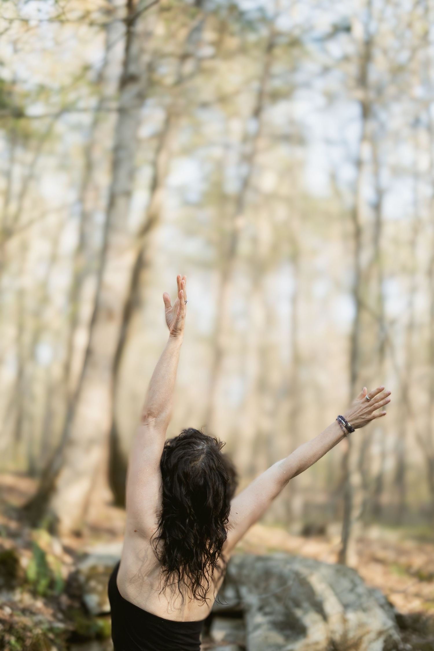 A person with dark wet hair raises their arms to the sky in a sunlit forest with tall trees and scattered rocks.