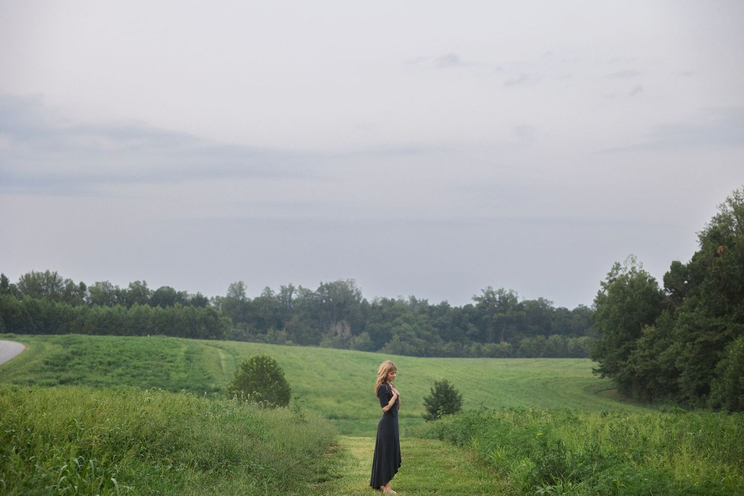 A woman in a dark dress stands on a grassy rural path surrounded by lush green fields and trees under a cloudy sky.
