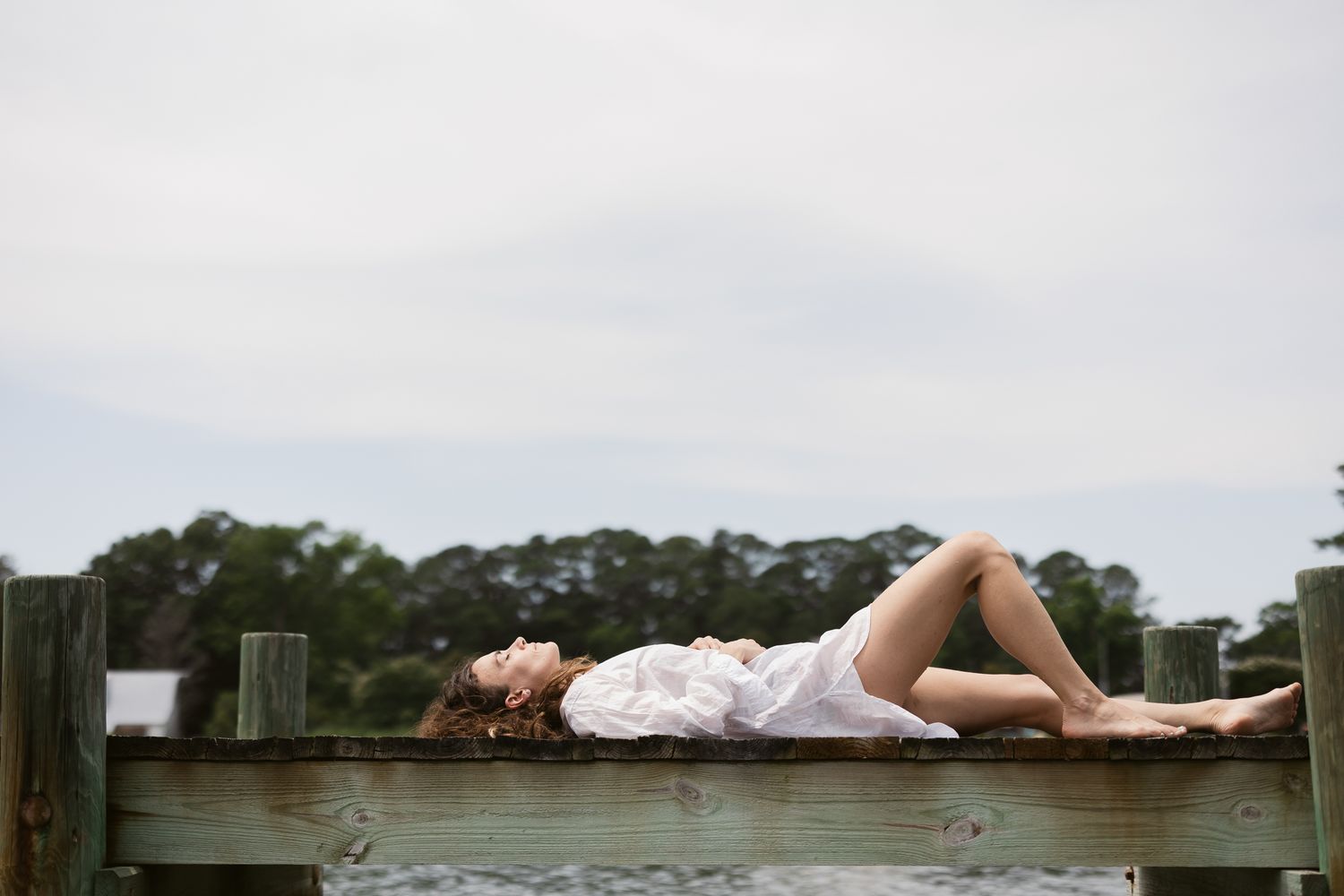 A person in white clothing lies peacefully on a wooden dock overlooking a serene lake surrounded by trees.