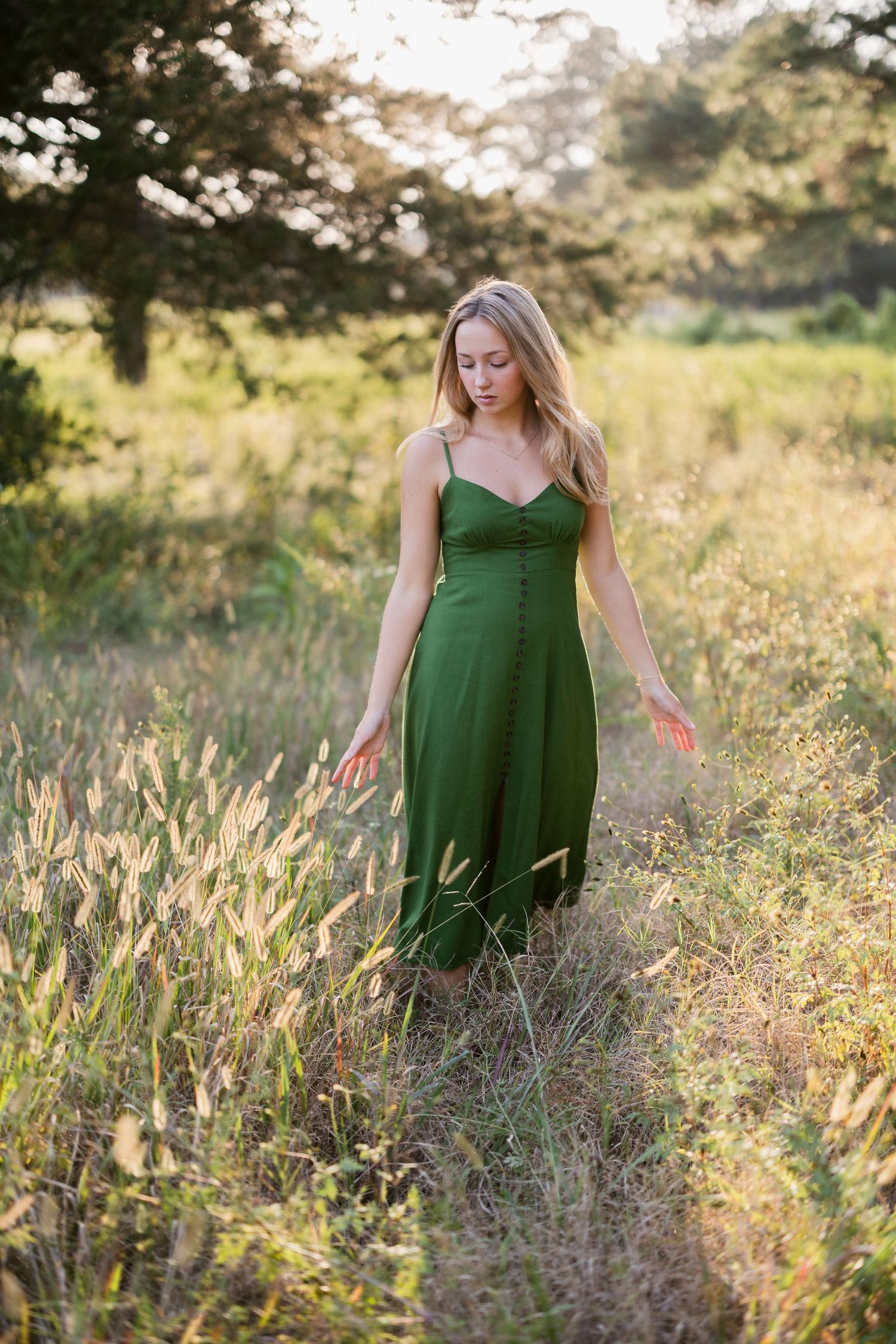 A woman in an emerald green dress poses gracefully among tall wildflowers in a sunlit meadow at golden hour.