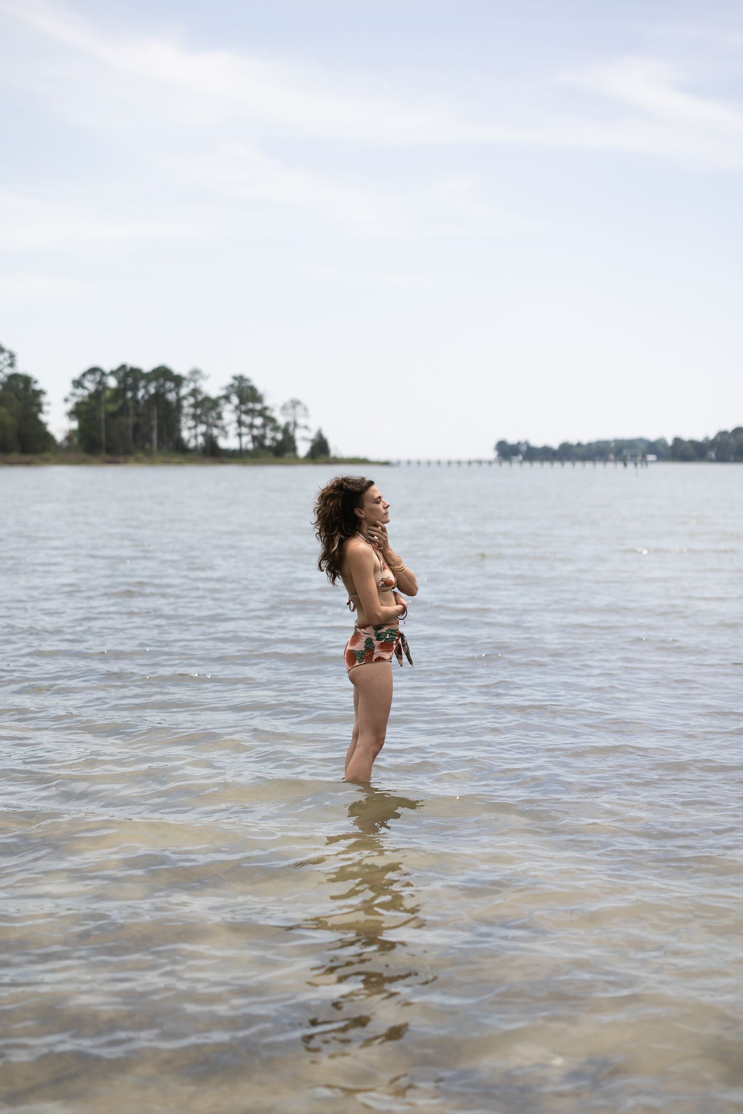 A person stands in shallow lake waters on an overcast day with trees and shoreline visible in the distance.