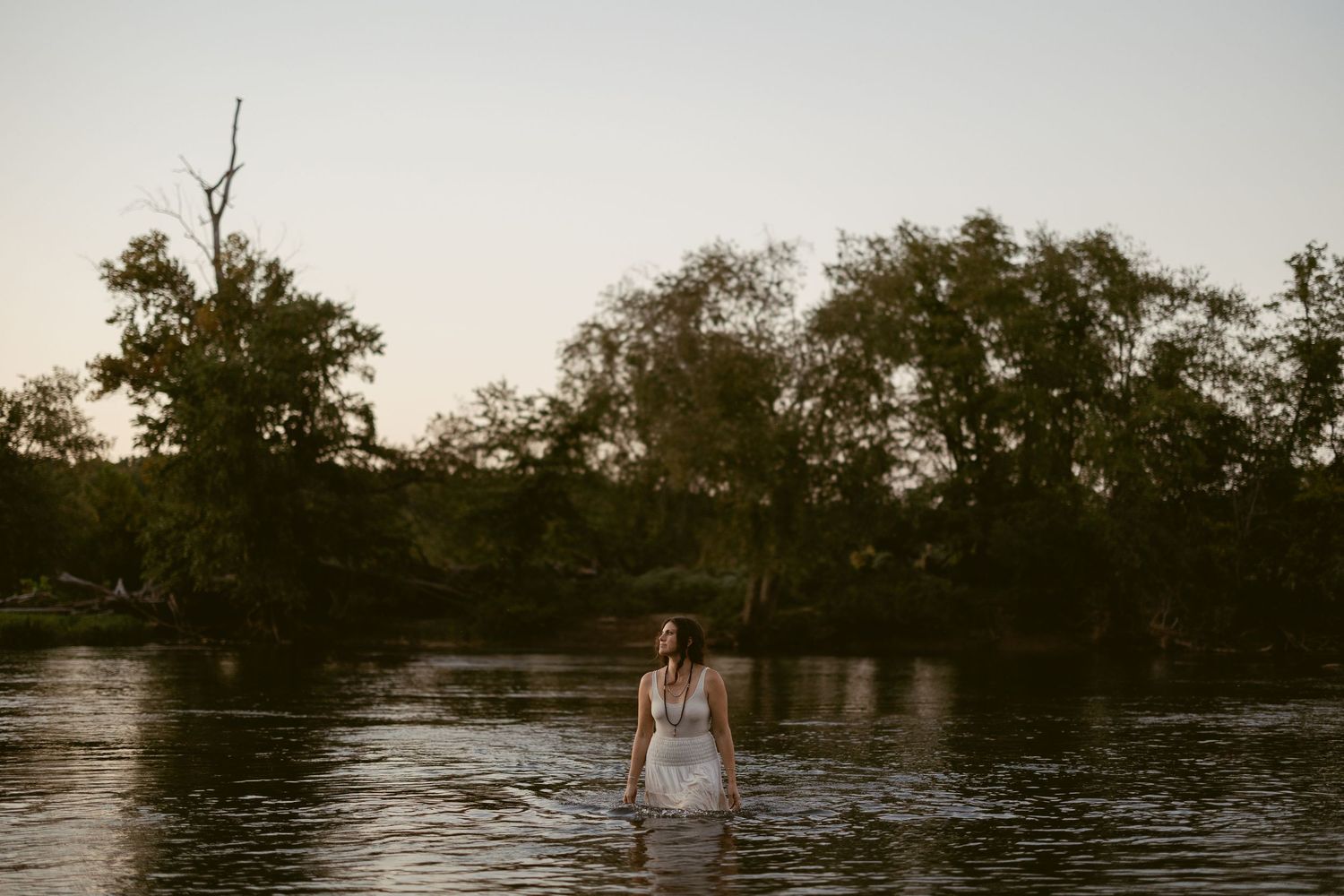 A scenic river landscape with trees and foliage along the shoreline at dusk creates a tranquil natural setting.