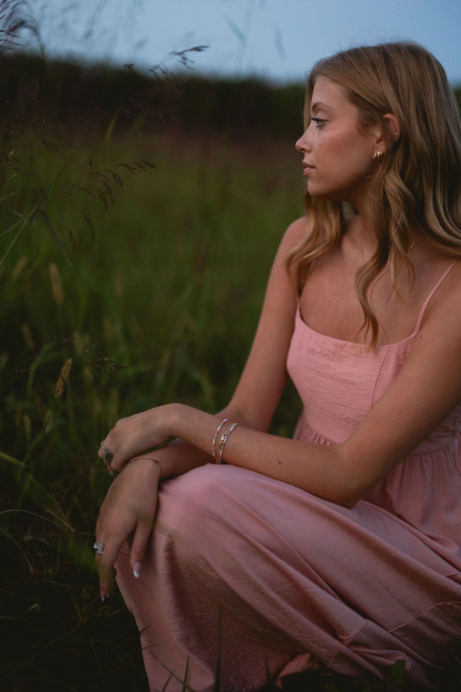 Young woman in pink dress crouching in a lush green field at dusk, wearing silver bracelets.