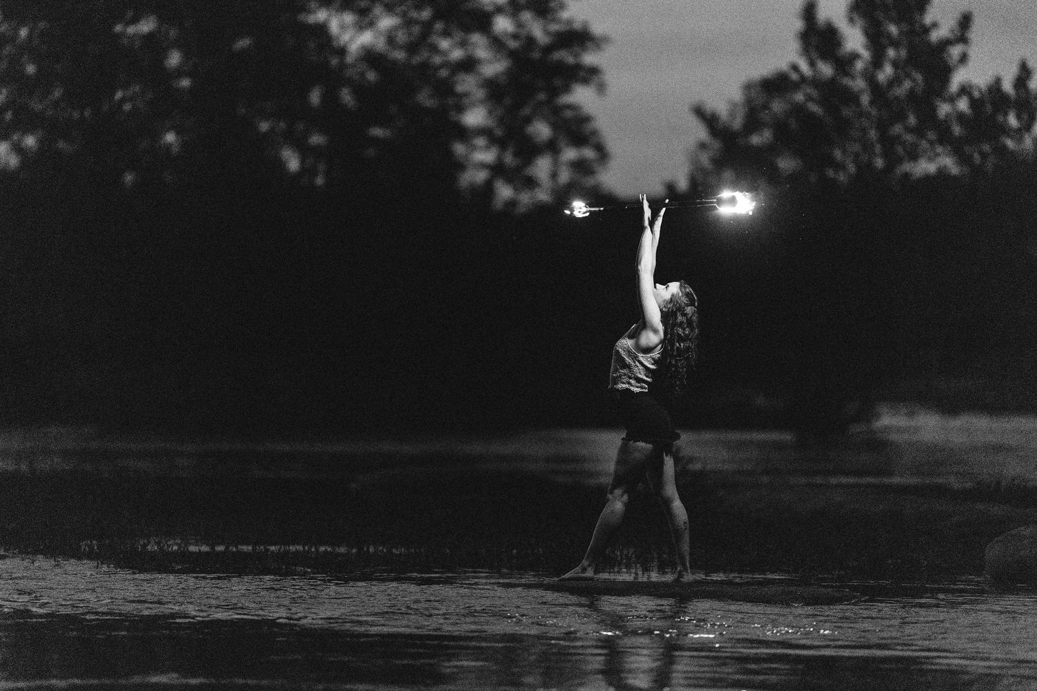 A silhouette jumps in a puddle at dusk with lights glowing in the background.
