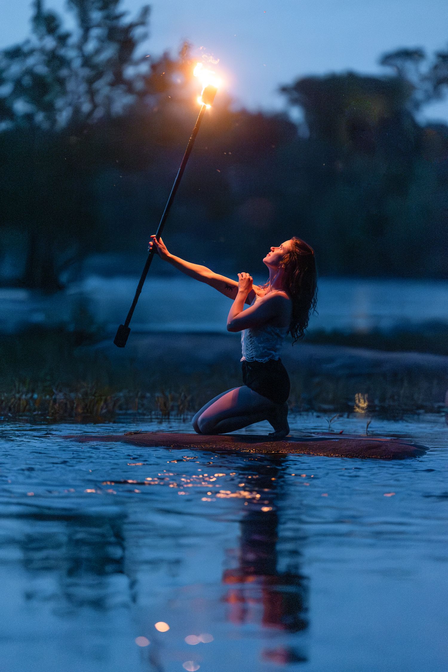Paddleboarder holding a glowing torch on calm waters at dusk while kneeling on the board.