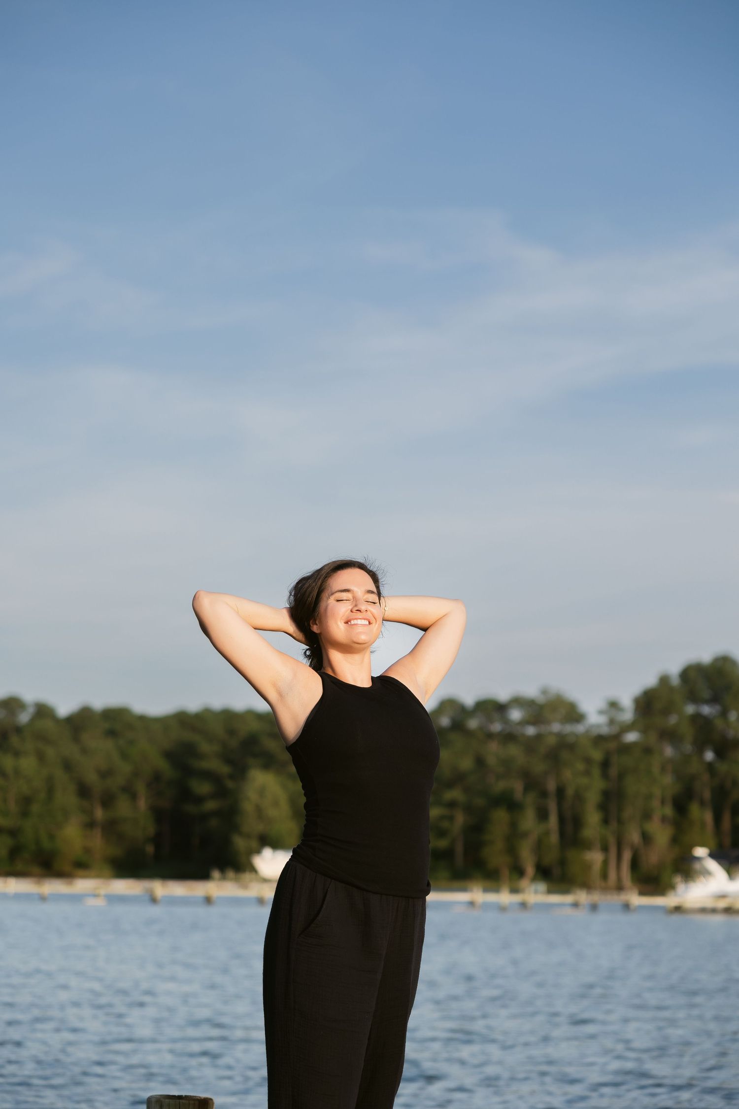 Person in black dress stretches peacefully by serene lake with trees and blue sky in background.