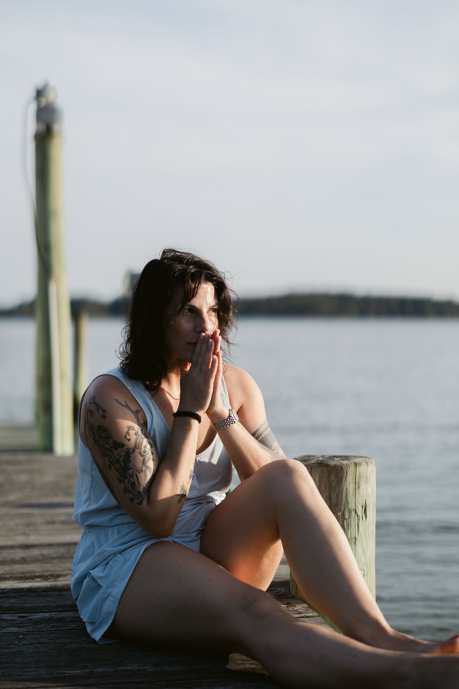 Someone sits thoughtfully on a wooden dock by the water wearing a light tank top on a calm summer evening.