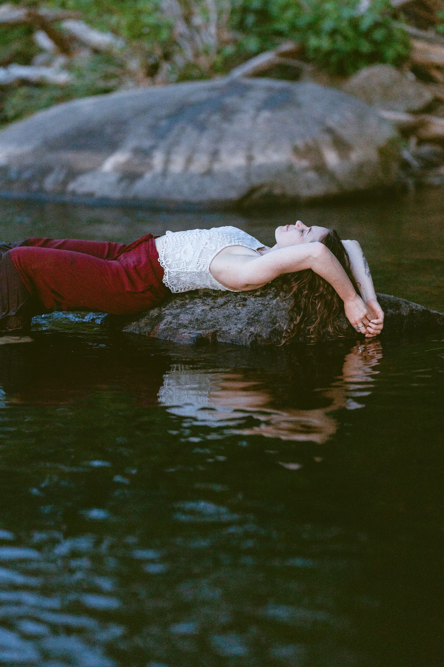 Person in red pants and white top floating gracefully on dark water near rocks in a natural setting.