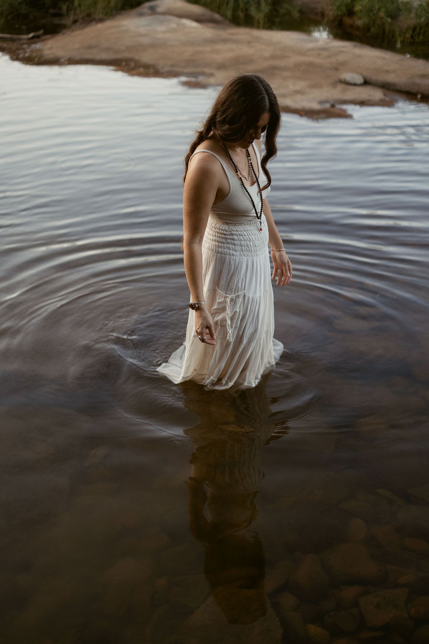 Person wearing a white dress wades into calm lake water at sunset creating rippling reflections.