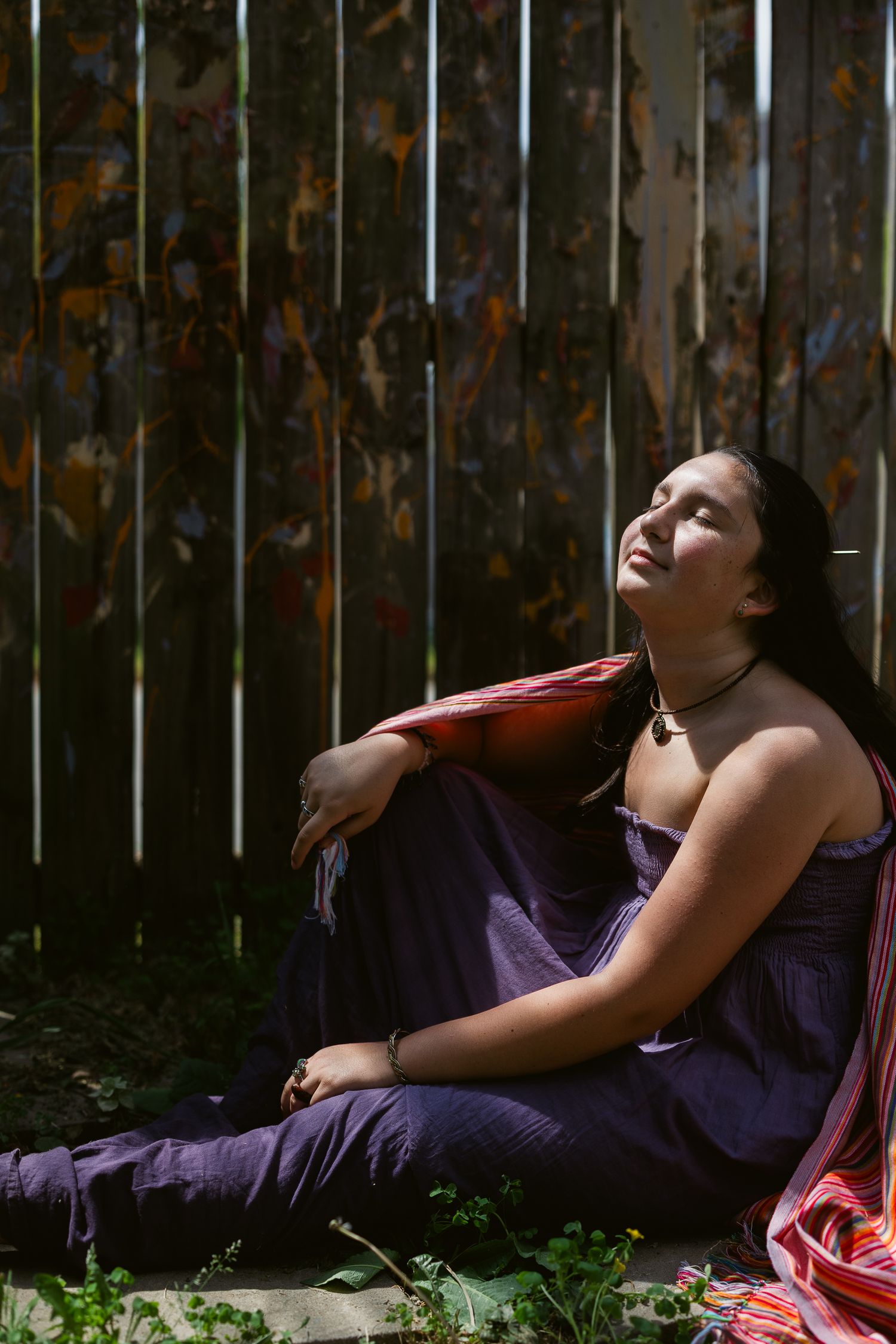 Young woman in purple dress sitting against a rustic wooden fence, gazing upward in natural sunlight.