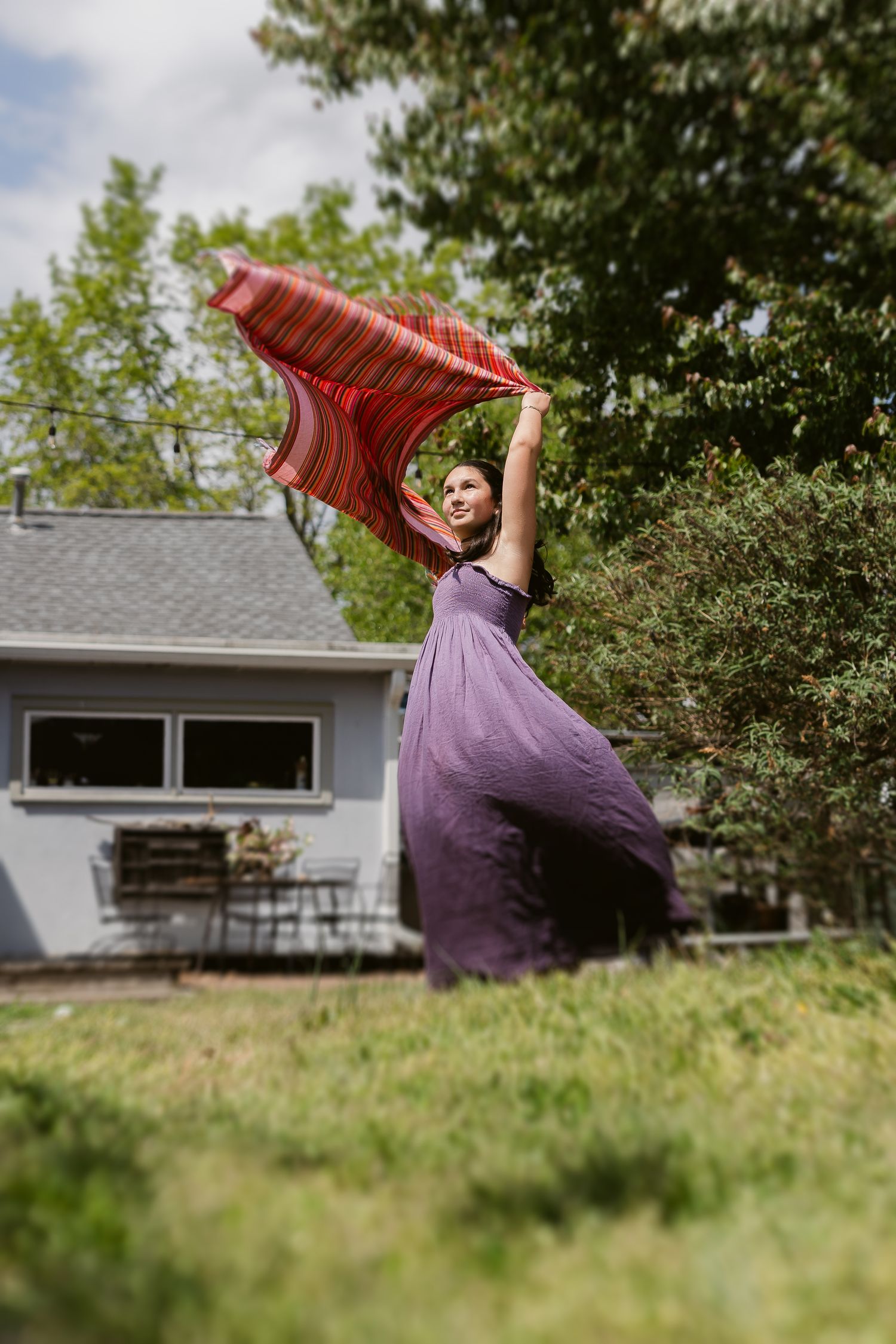 Woman in purple maxi dress twirling red striped scarf in sunny backyard with green trees and house.